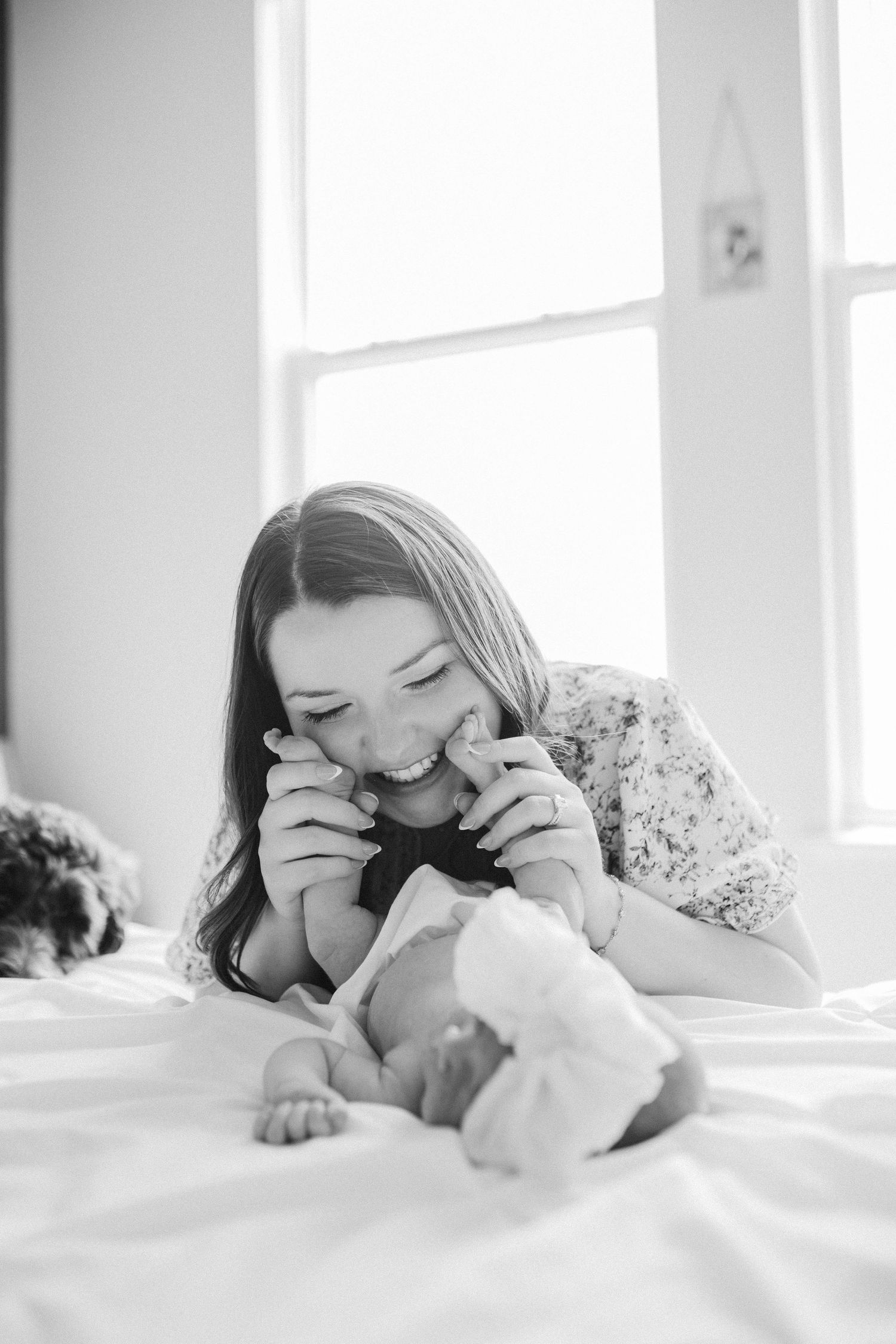 Black and white photo of a smiling mother playfully kissing her newborn baby's hands while lying on a white bed.