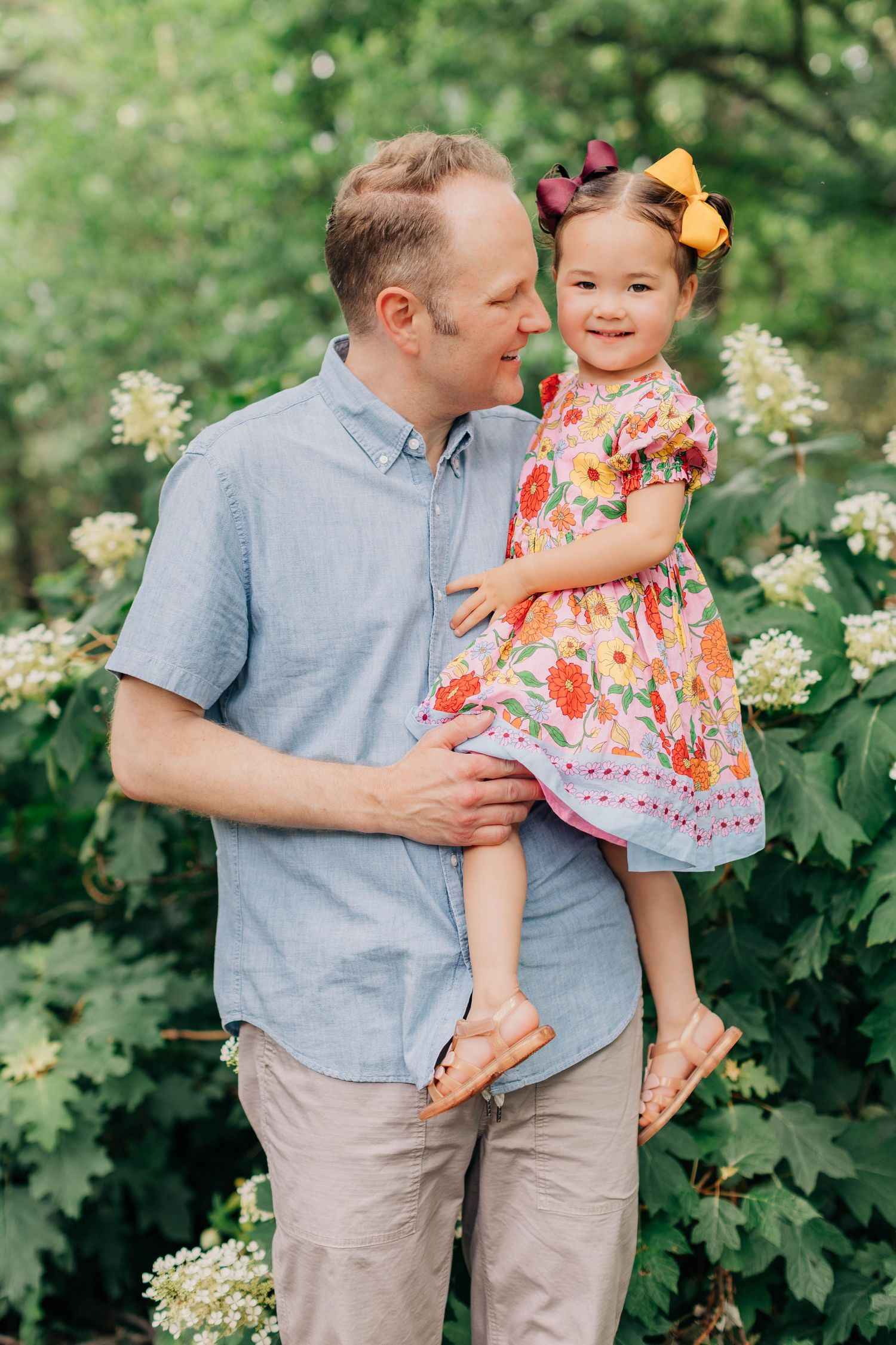 A father holds his smiling young daughter in a floral dress with colorful hair bows among white blooming flowers outdoors.