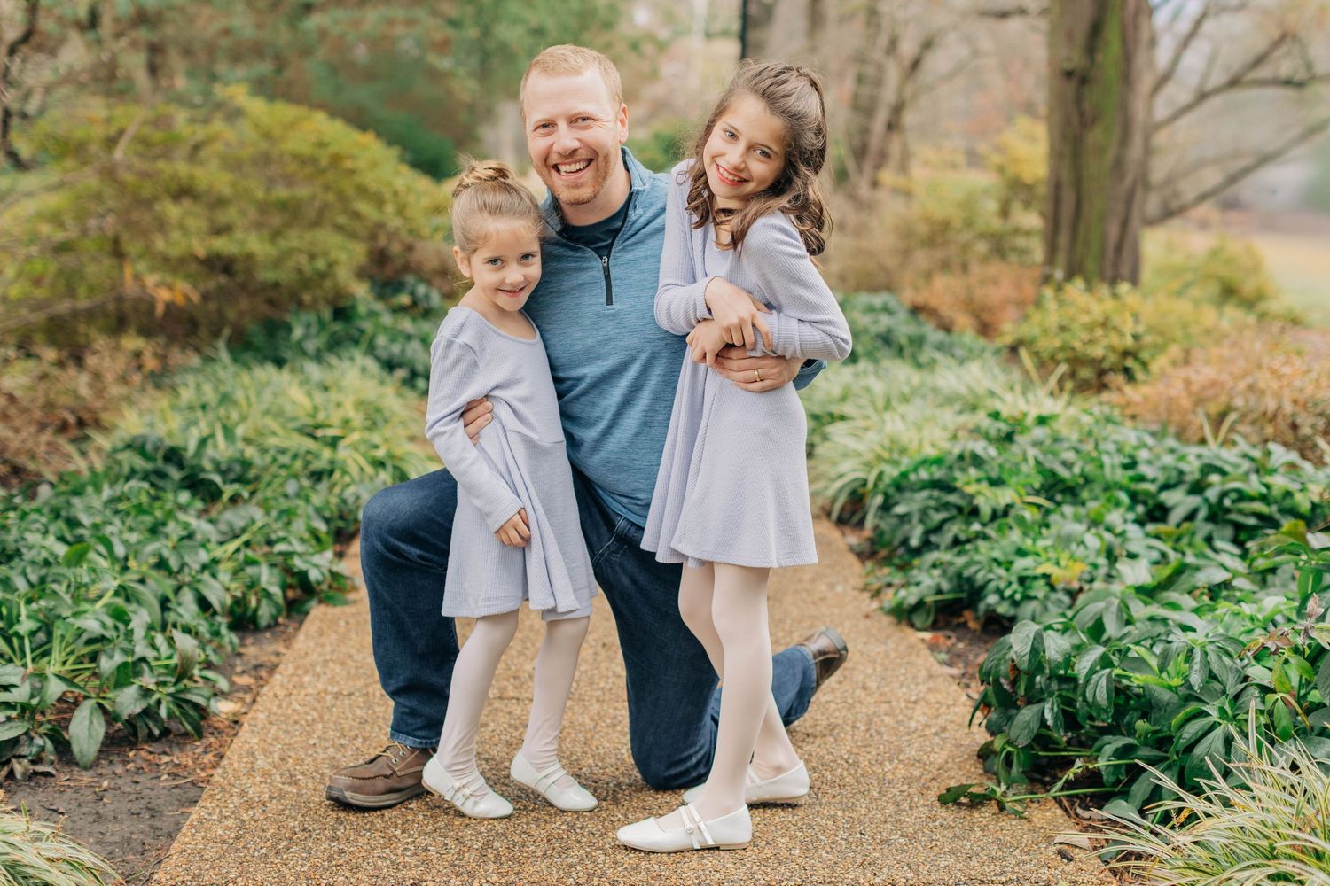 A family of three poses together on a garden path surrounded by lush greenery.