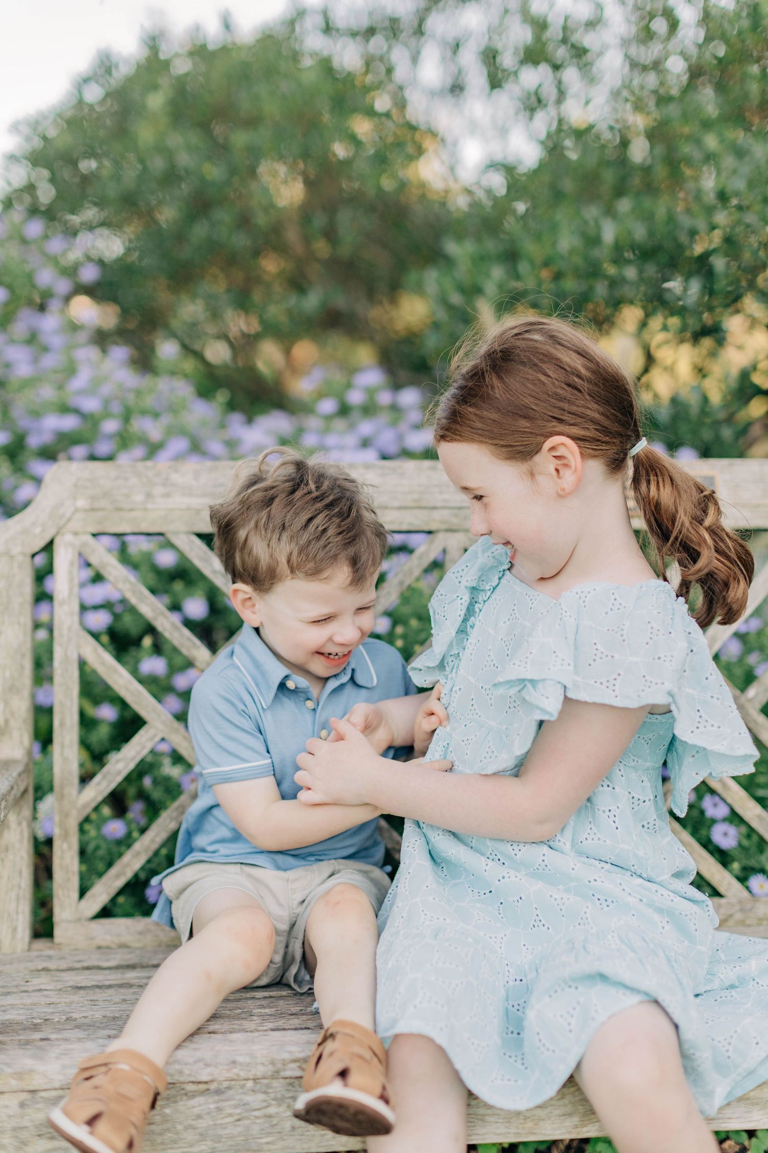 Two children share a playful moment while sitting on a wooden bench in a flower garden.