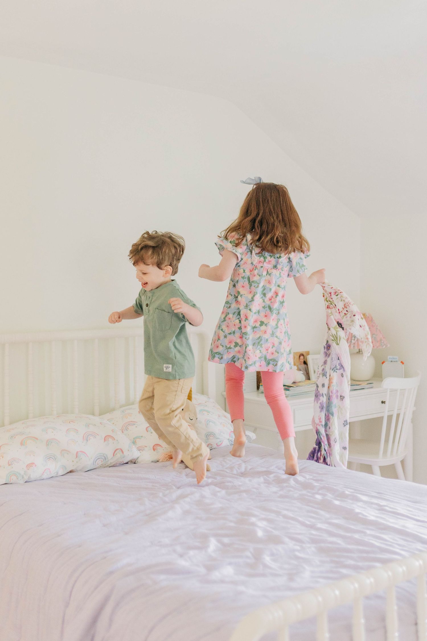 Children having fun playing and bouncing on a white bed in a bright room.