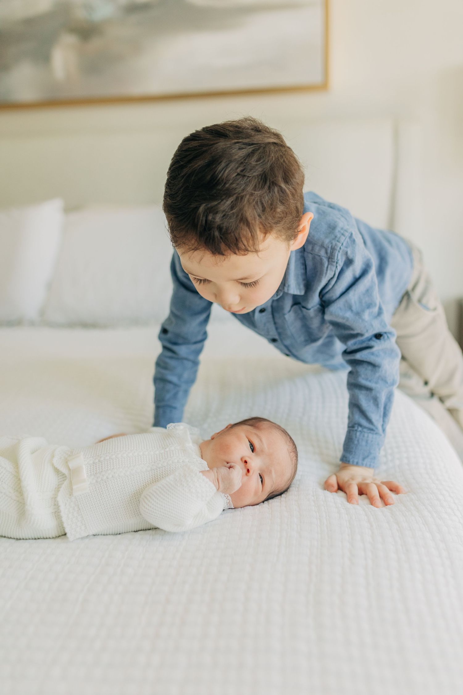 A child in a blue sweater leans over to look at a baby on a white bed.