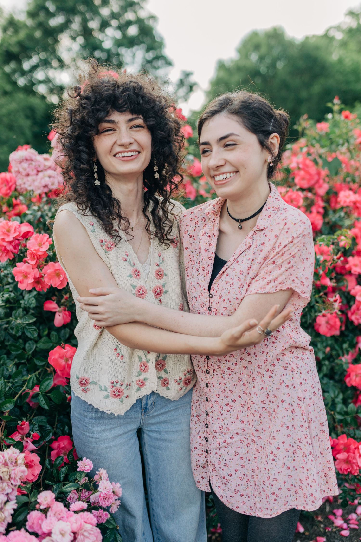 Two friends share a candid moment of laughter among vibrant pink rose bushes on a sunny day.