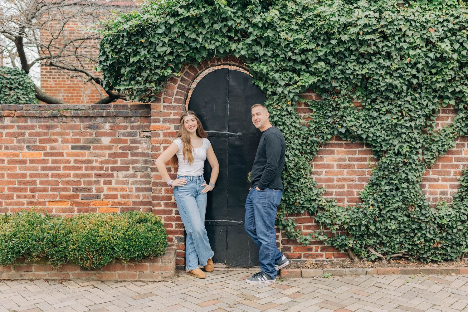 A couple standing by an ivy-covered brick wall with a black door in casual attire.