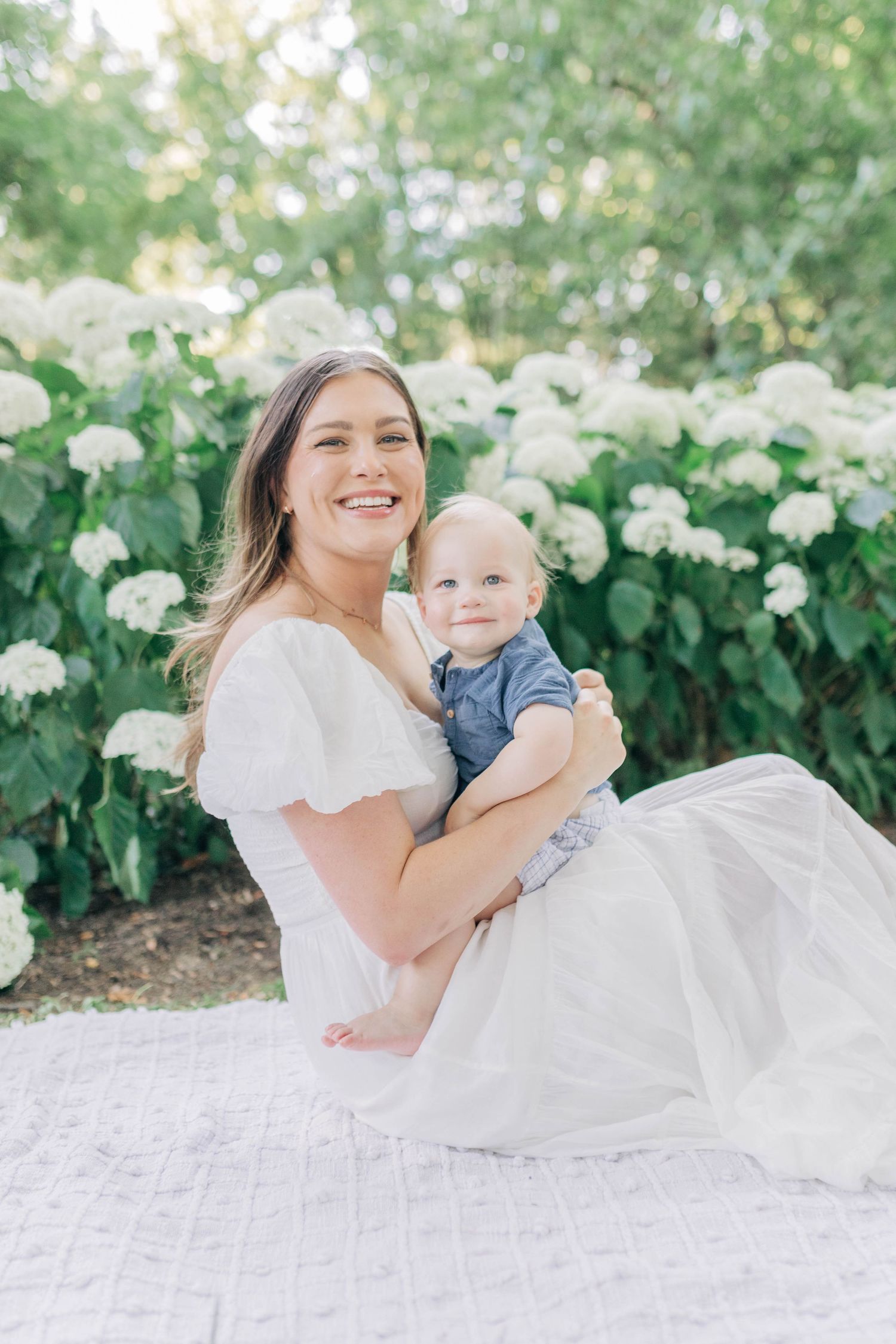 Mother and baby share joyful moment on white blanket surrounded by blooming flowers.