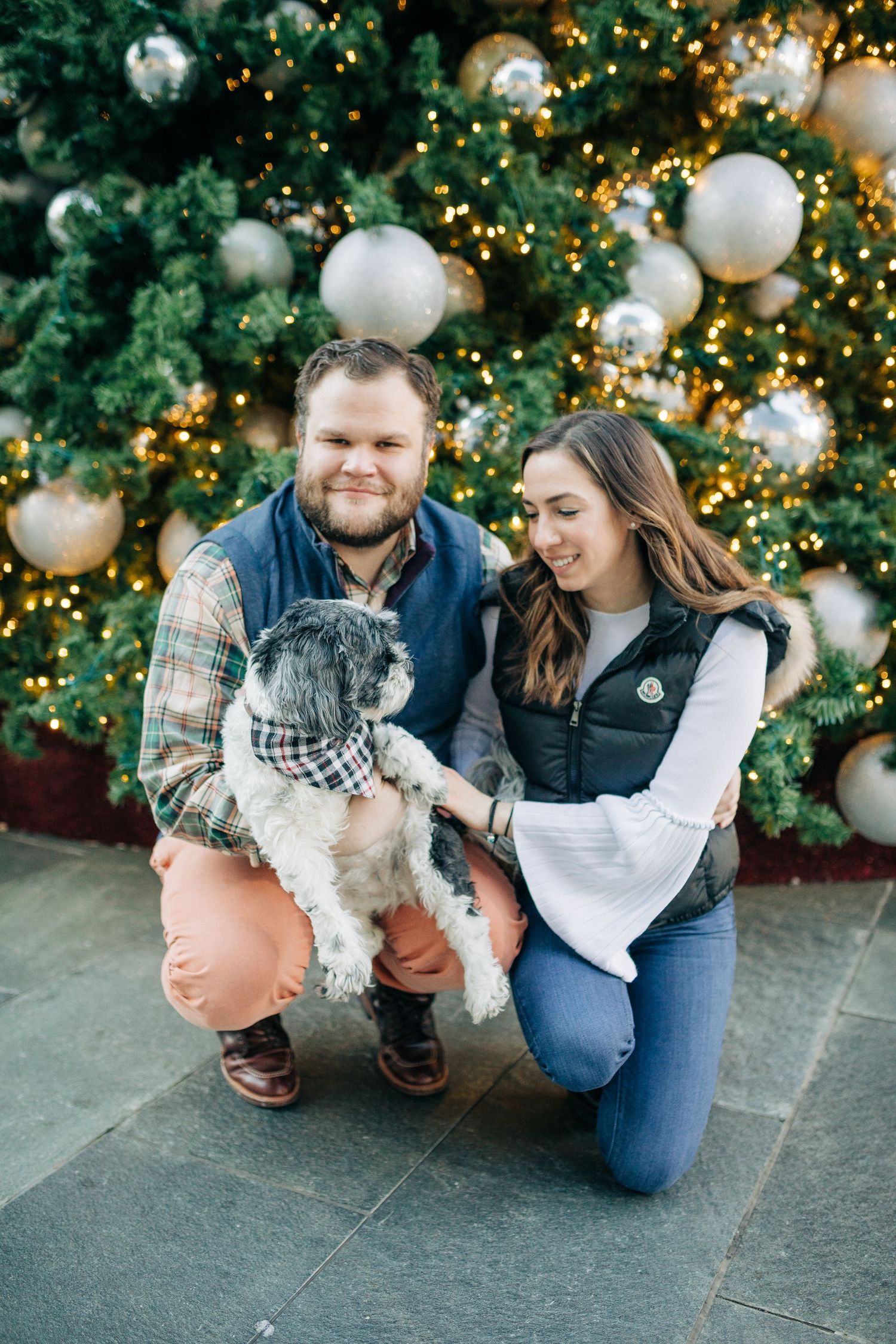 A couple kneels with their small dog in front of a decorated Christmas tree.