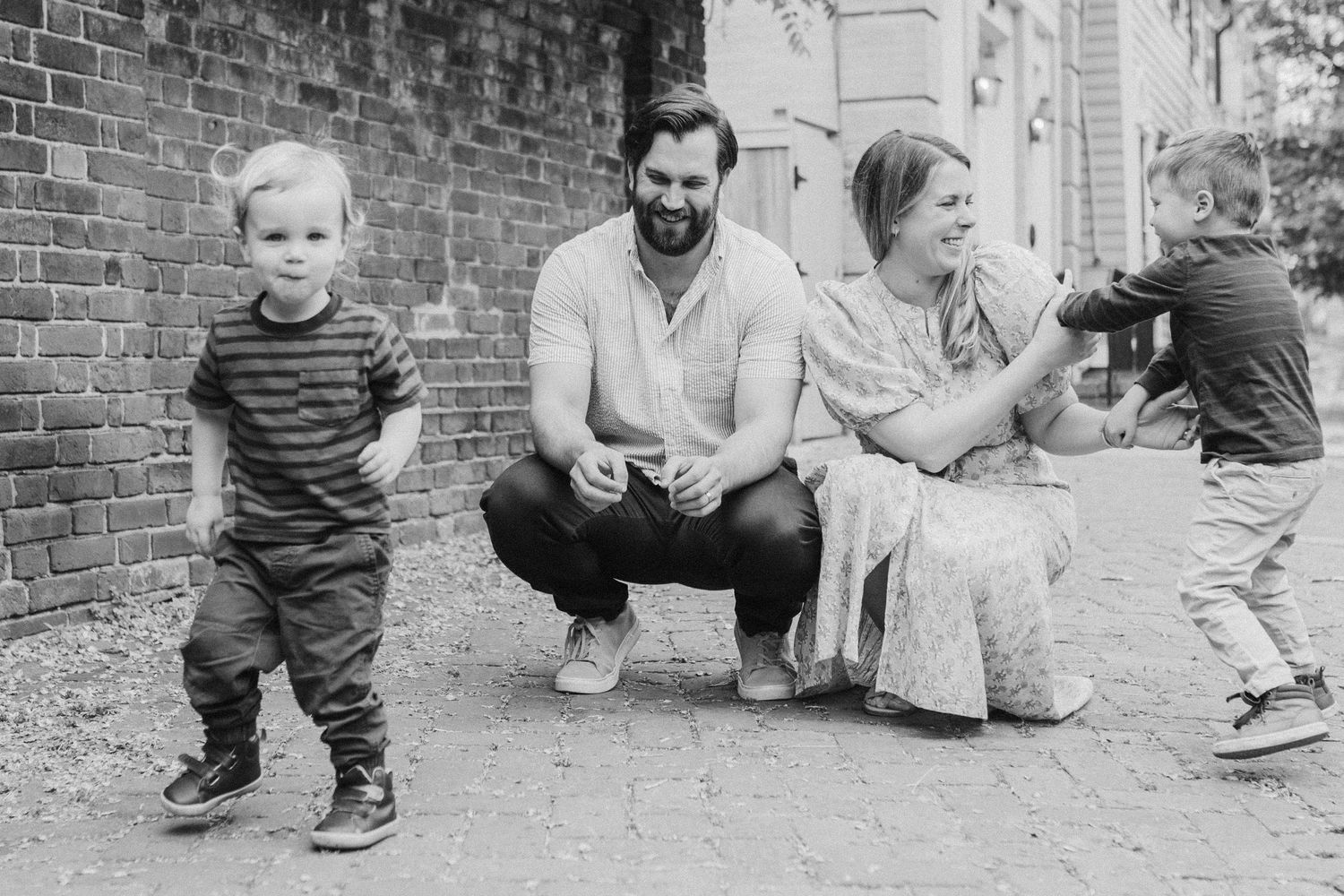 Family members crouch together against brick wall for casual black and white portrait.
