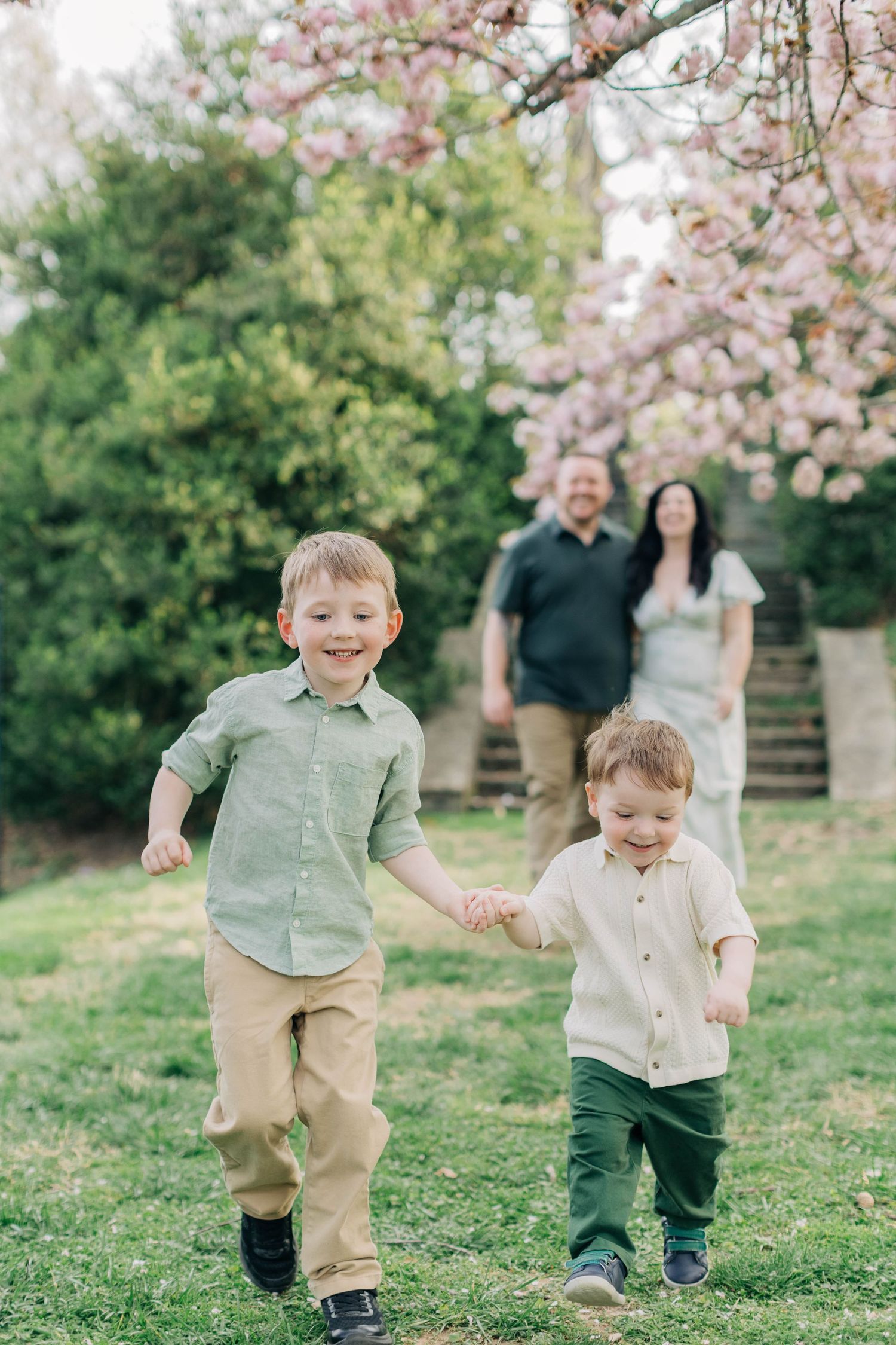 Two young brothers hold hands running in a spring garden while their parents smile in the background near pink blossoms.