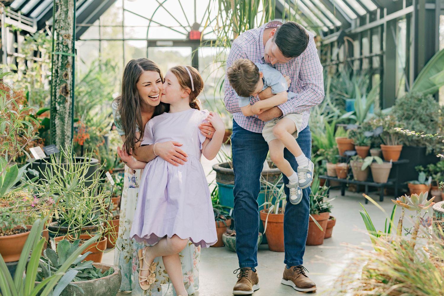 Family of four enjoying a stroll in a lush greenhouse, parents hugging children among potted plants.