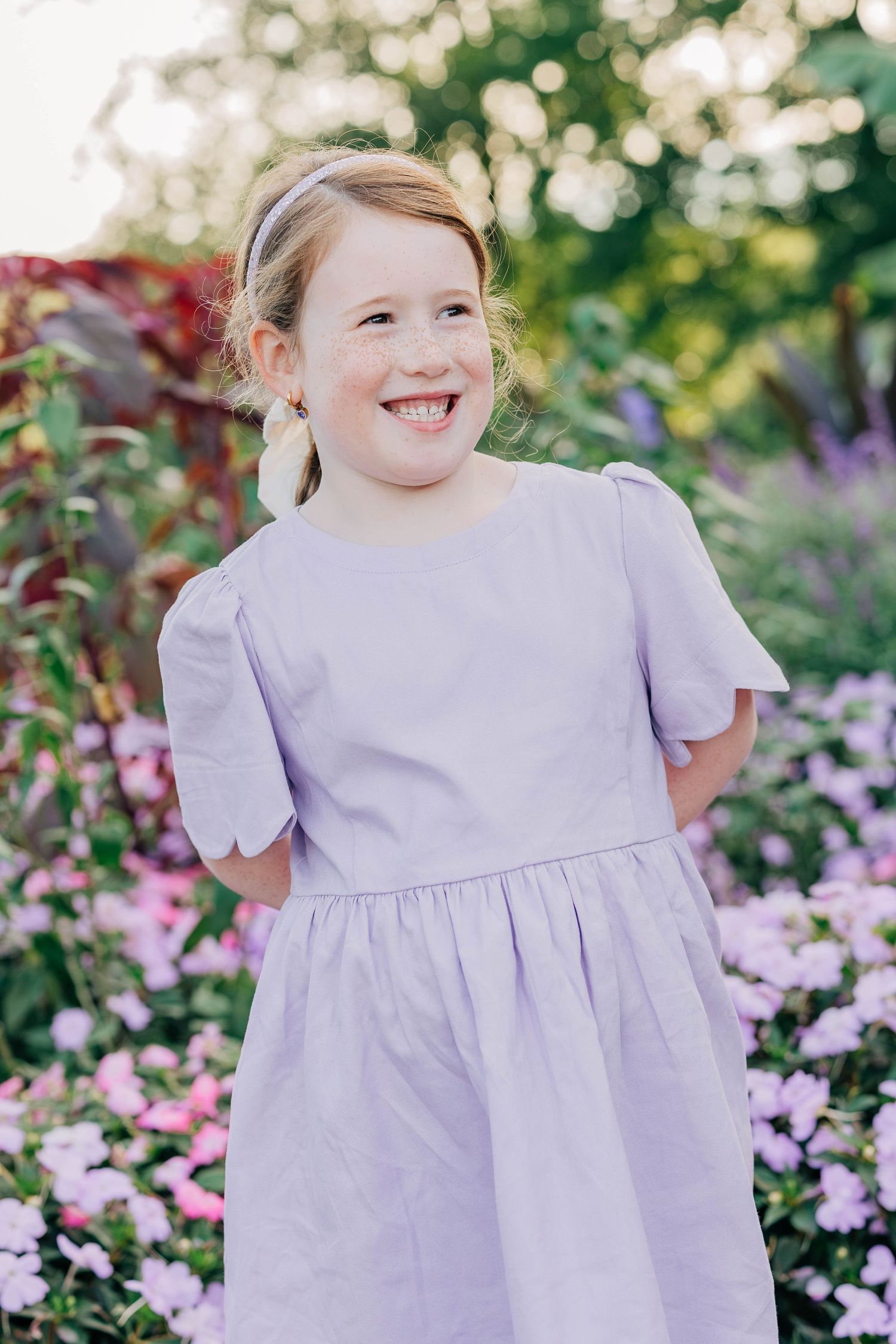 A joyful young girl in a lavender dress smiles among pink flowers in a garden.