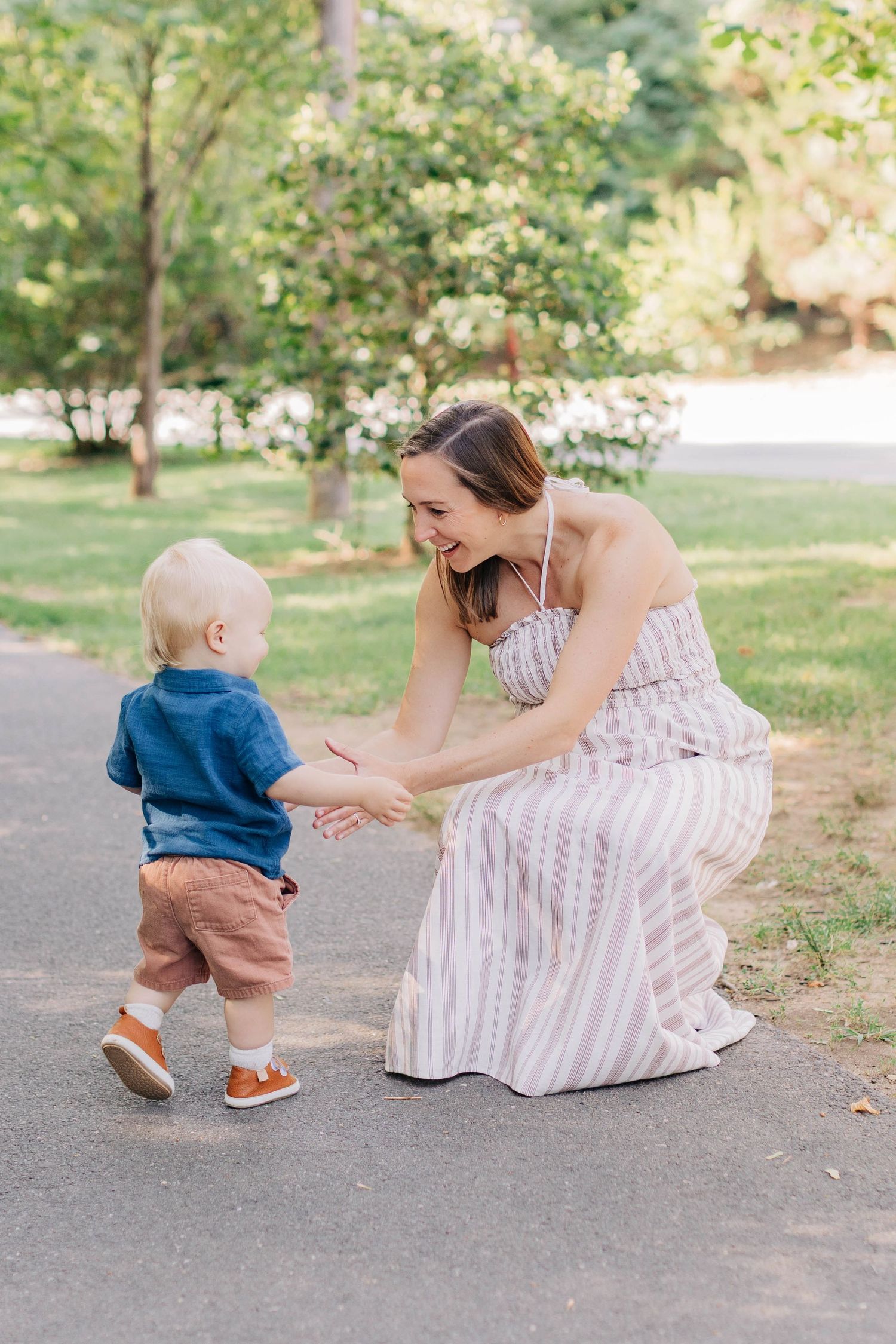 A smiling woman in a striped dress crouches down to hold hands with a toddler boy on a sunny park path.