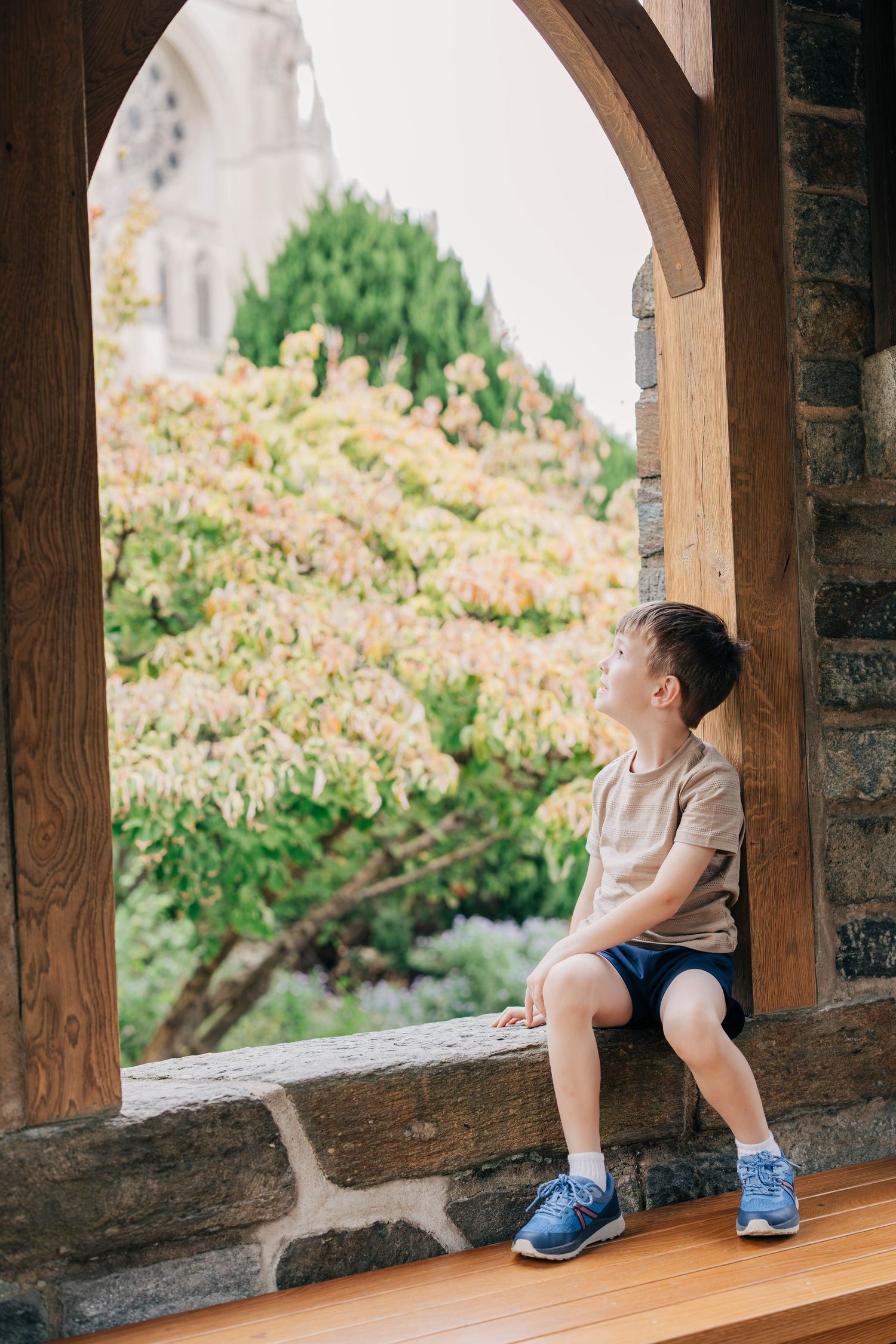 Young boy in a tan t-shirt and blue shorts sits on a stone ledge in a wooden archway, gazing at a colorful tree and a distant cathedral.