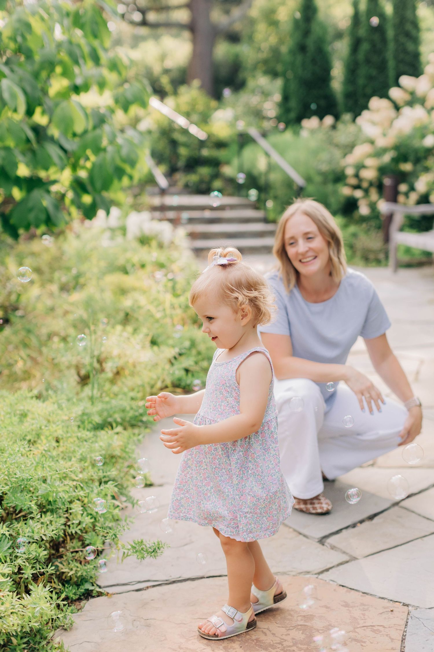 A smiling blonde woman crouches behind a toddler girl in a floral dress chasing soap bubbles in a lush green garden.
