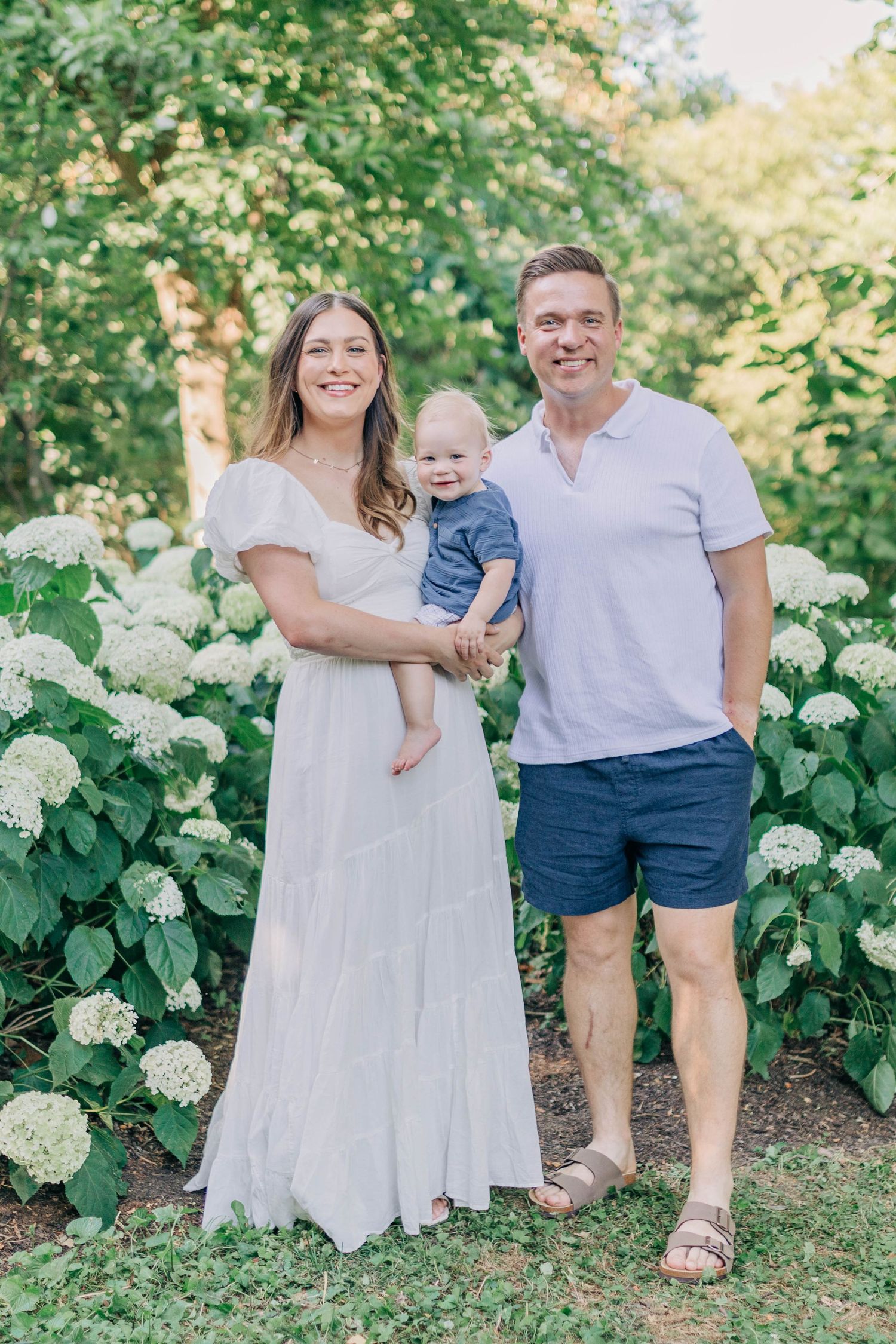 Family dressed in white and blue poses together in a summer garden with hydrangeas.