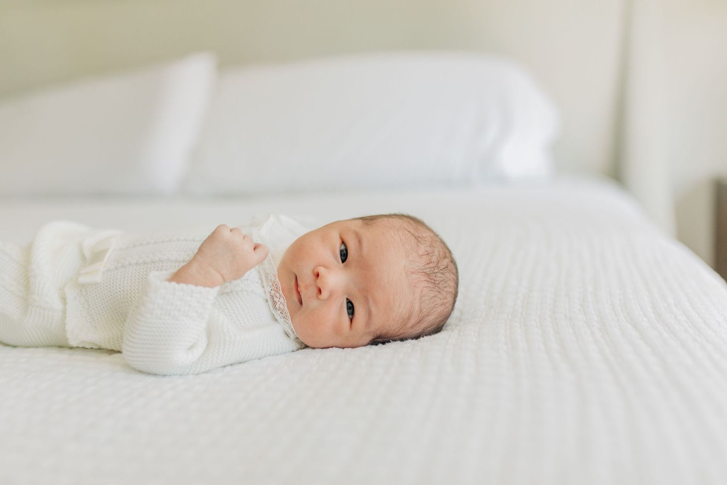Newborn baby wearing white sleepwear lying on white bedding in soft natural light.
