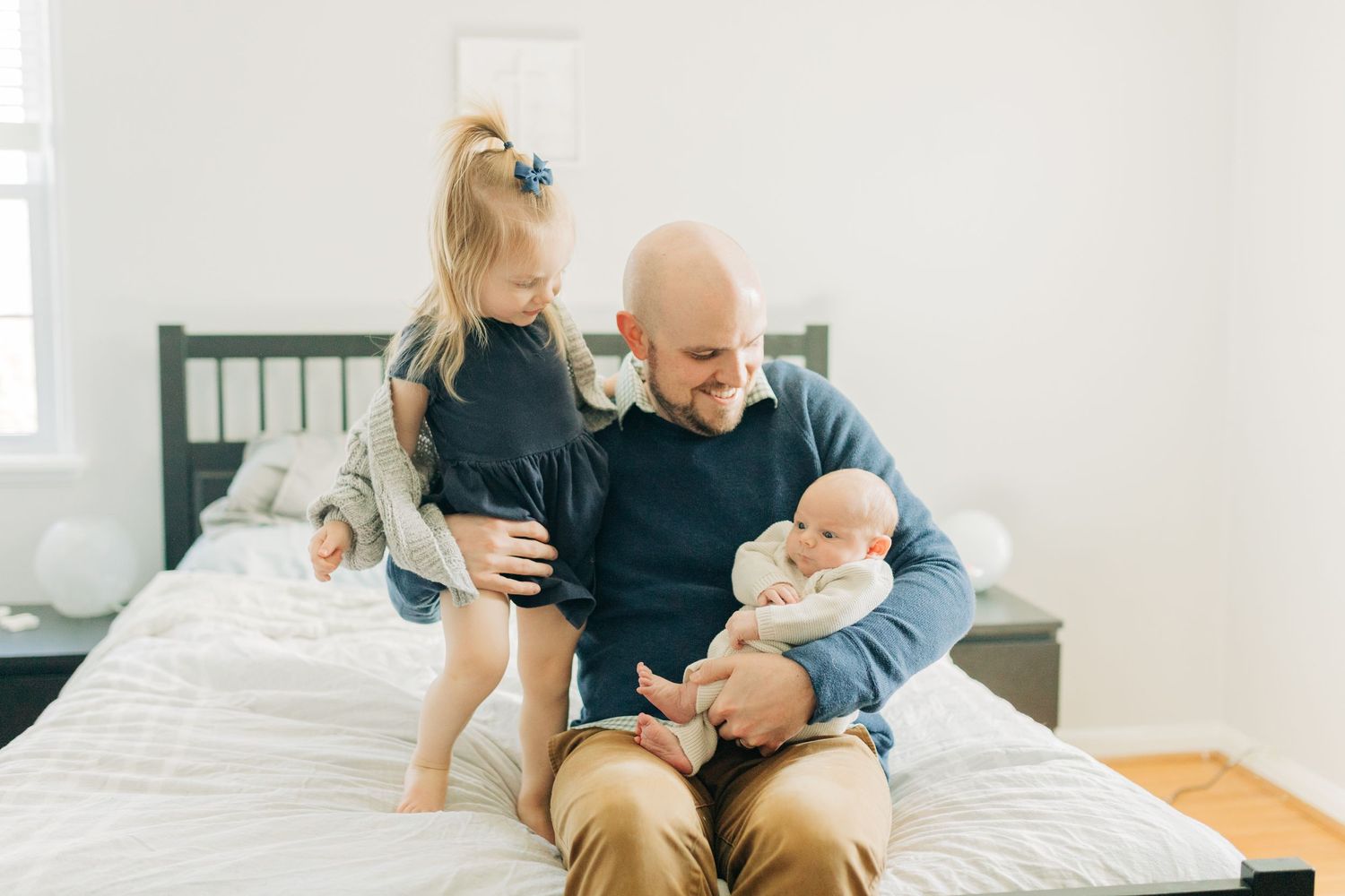Tender family moment captured in soft light as parents cuddle with their newborn on a white bed.