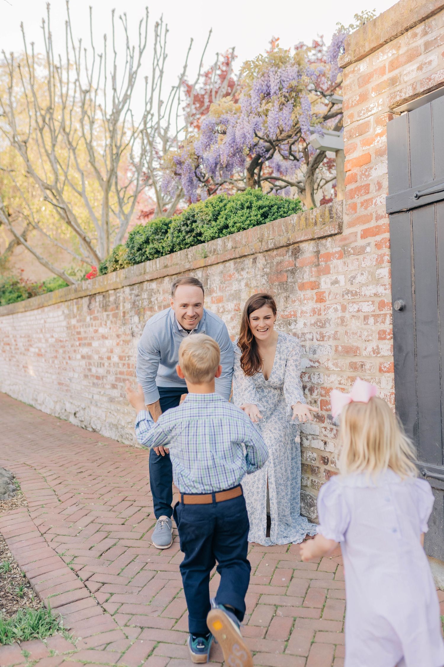 Family with two young children reuniting on a brick pathway beside a wisteria-covered wall in spring.
