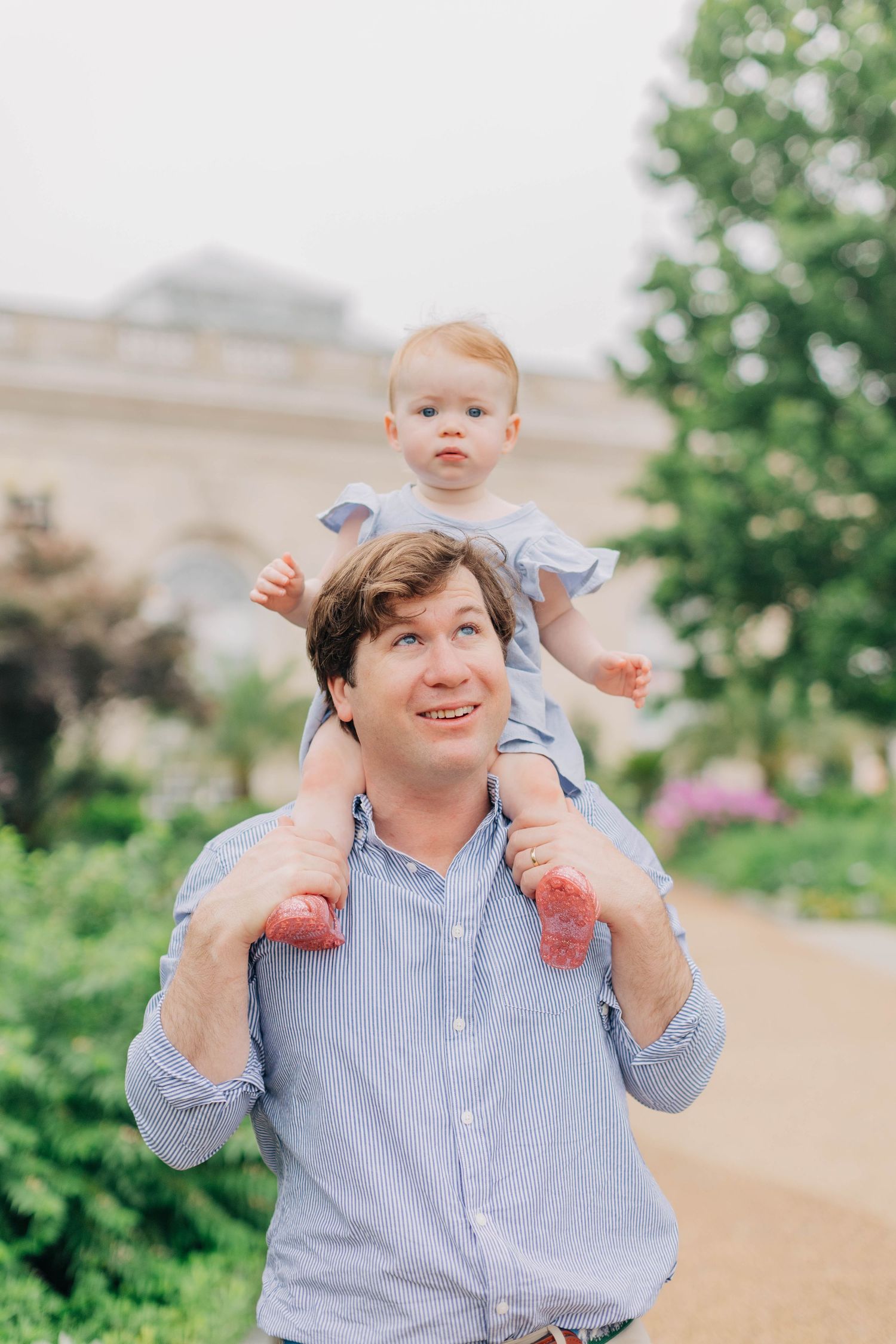 A smiling father carries his baby girl on his shoulders outdoors in a lush green park setting.
