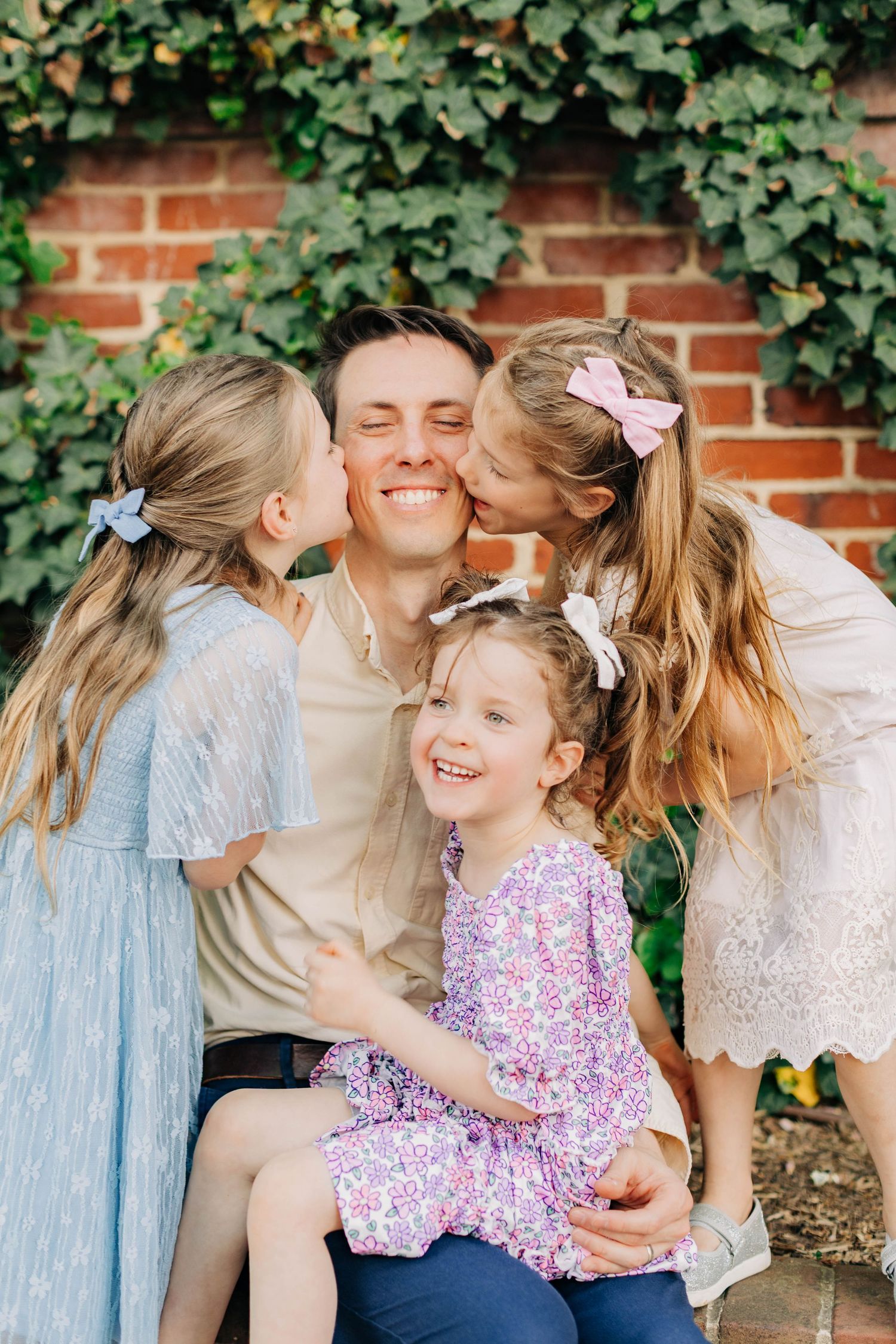 A happy father sits with his three young daughters, two kissing his cheeks while one smiles in his lap.