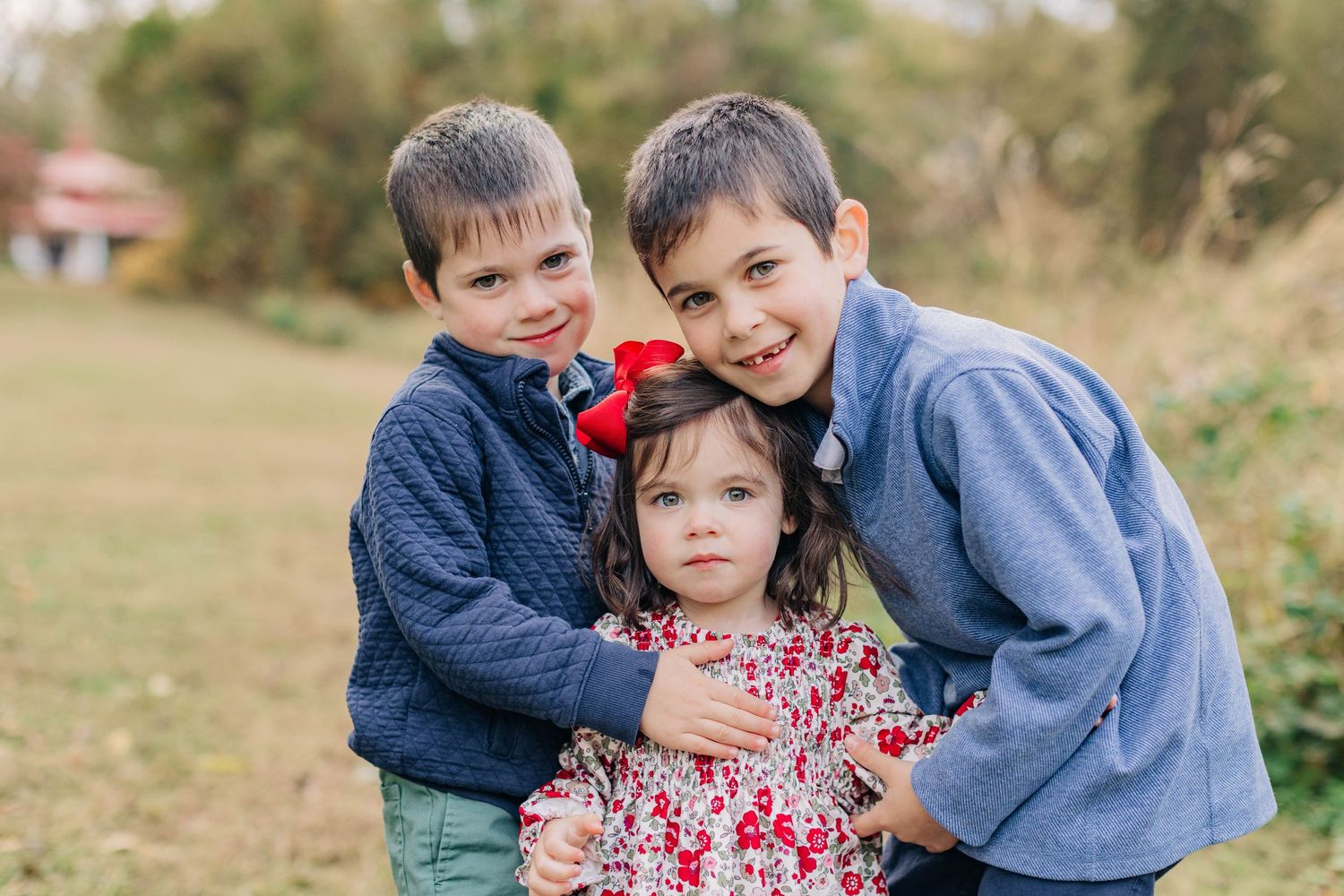 Three young siblings pose outdoors, two boys hugging a little girl in a floral dress with a red bow.