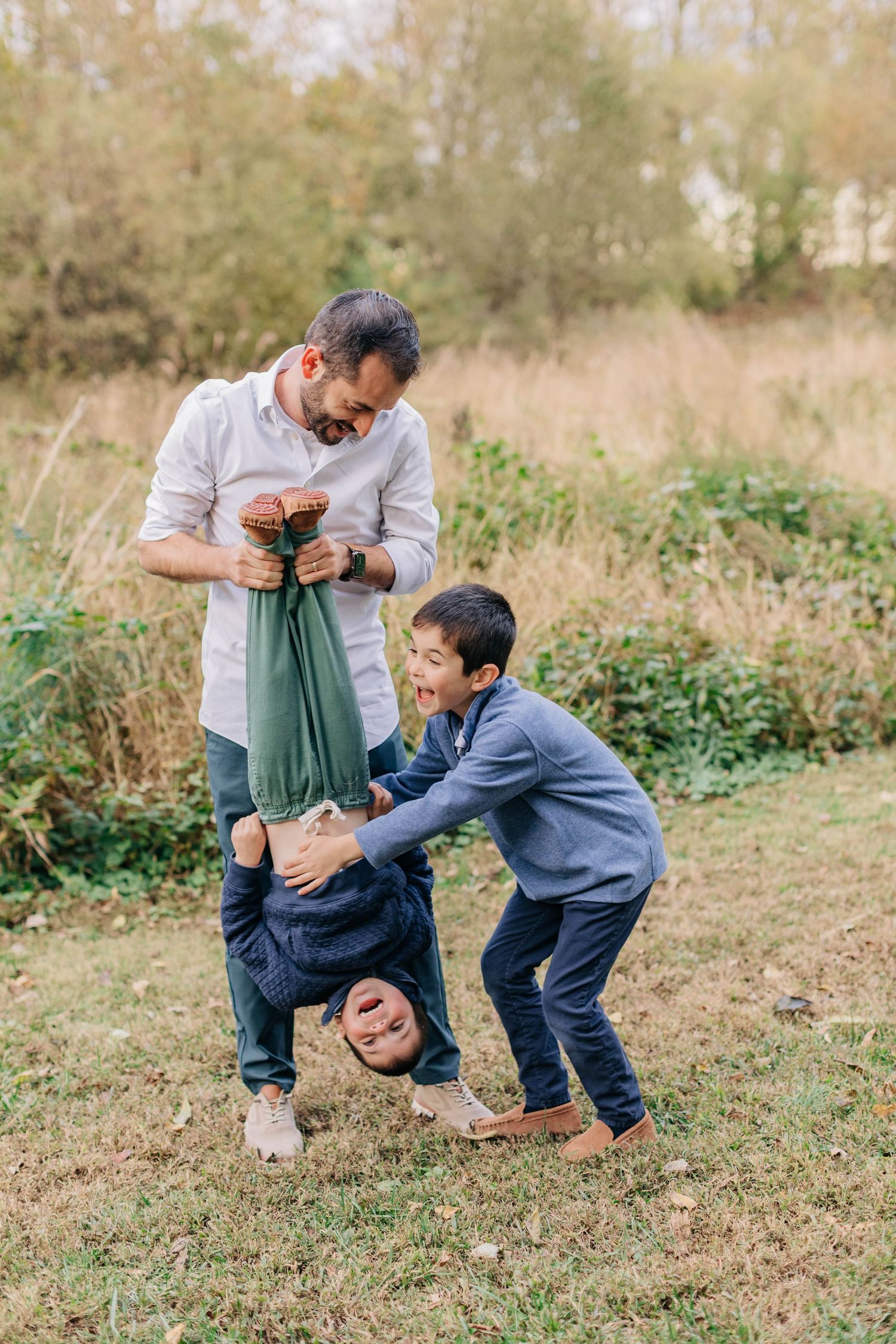 A father playfully holds a child upside down while another boy laughs outdoors in a grassy field.
