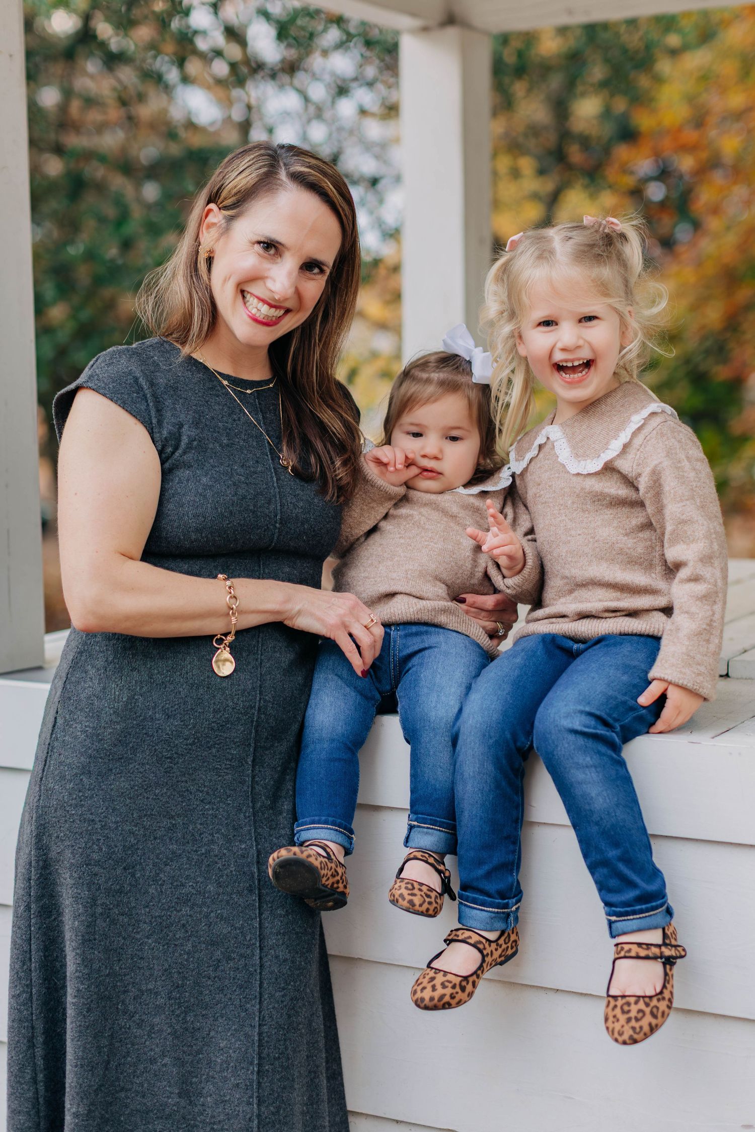 A smiling woman in a gray dress poses with two young girls in matching tan sweaters and jeans on a porch.
