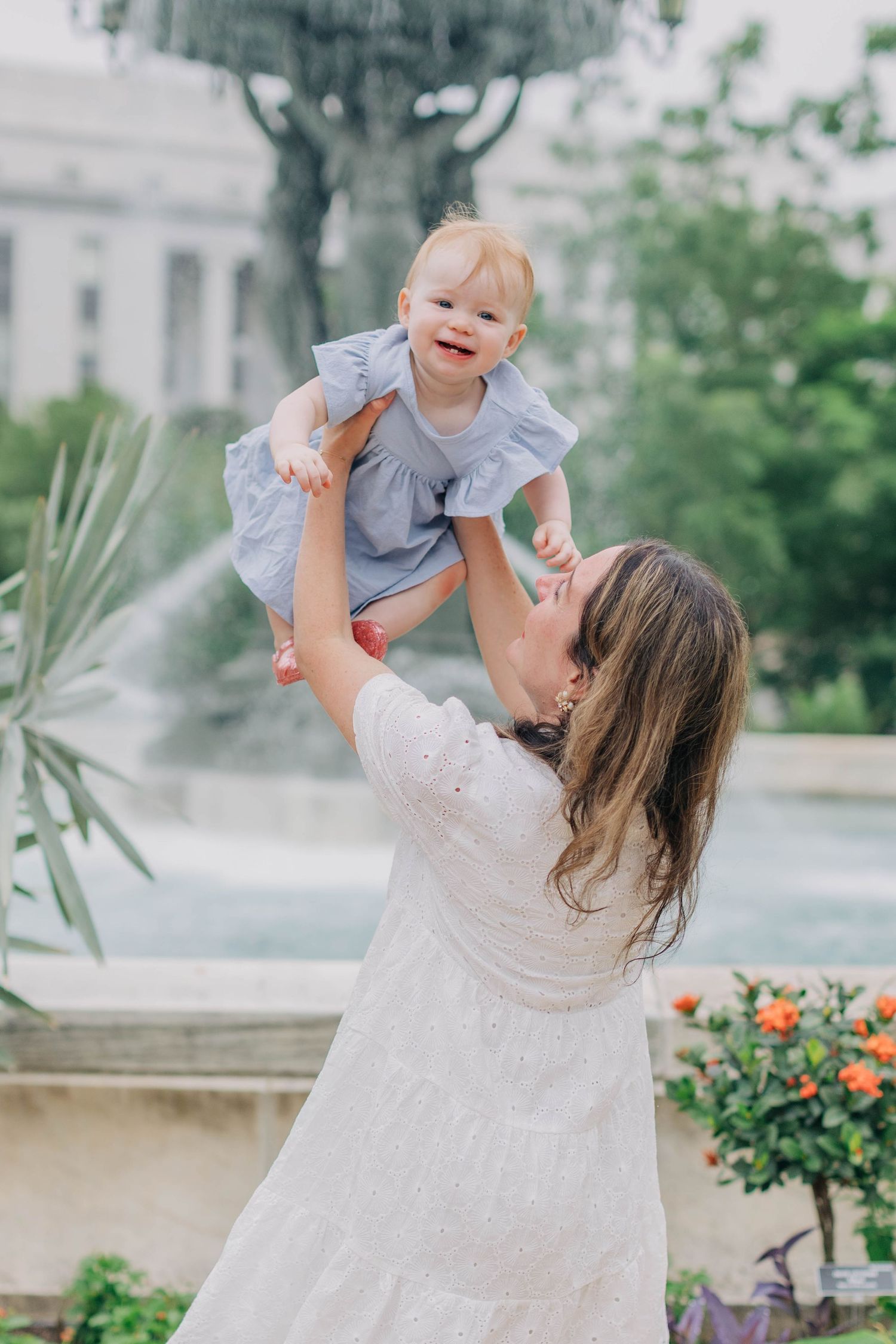 Mother lifting smiling baby girl in a blue dress near a fountain in a lush garden setting.