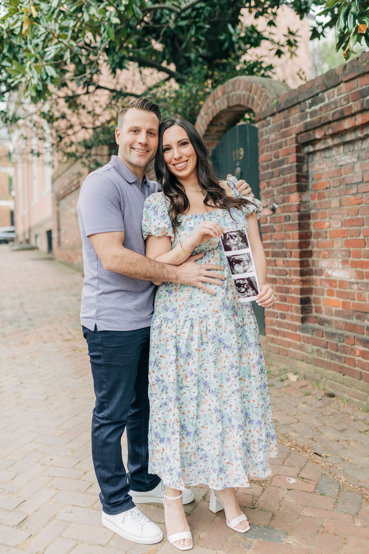 A couple celebrates while displaying ultrasound photos on a charming cobblestone street.