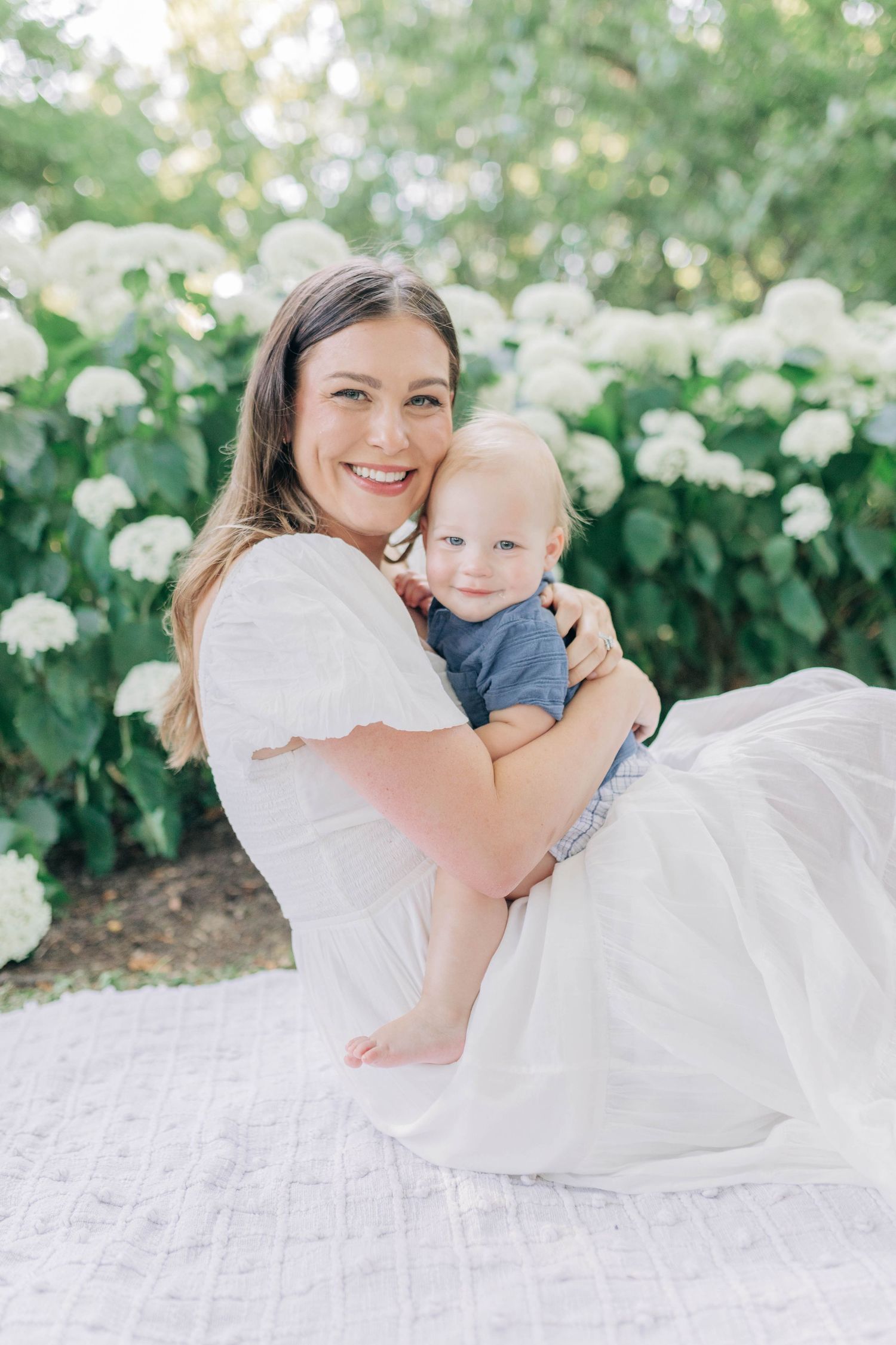 Mother and baby share joyful moments on white blanket surrounded by blooming flowers in garden setting.
