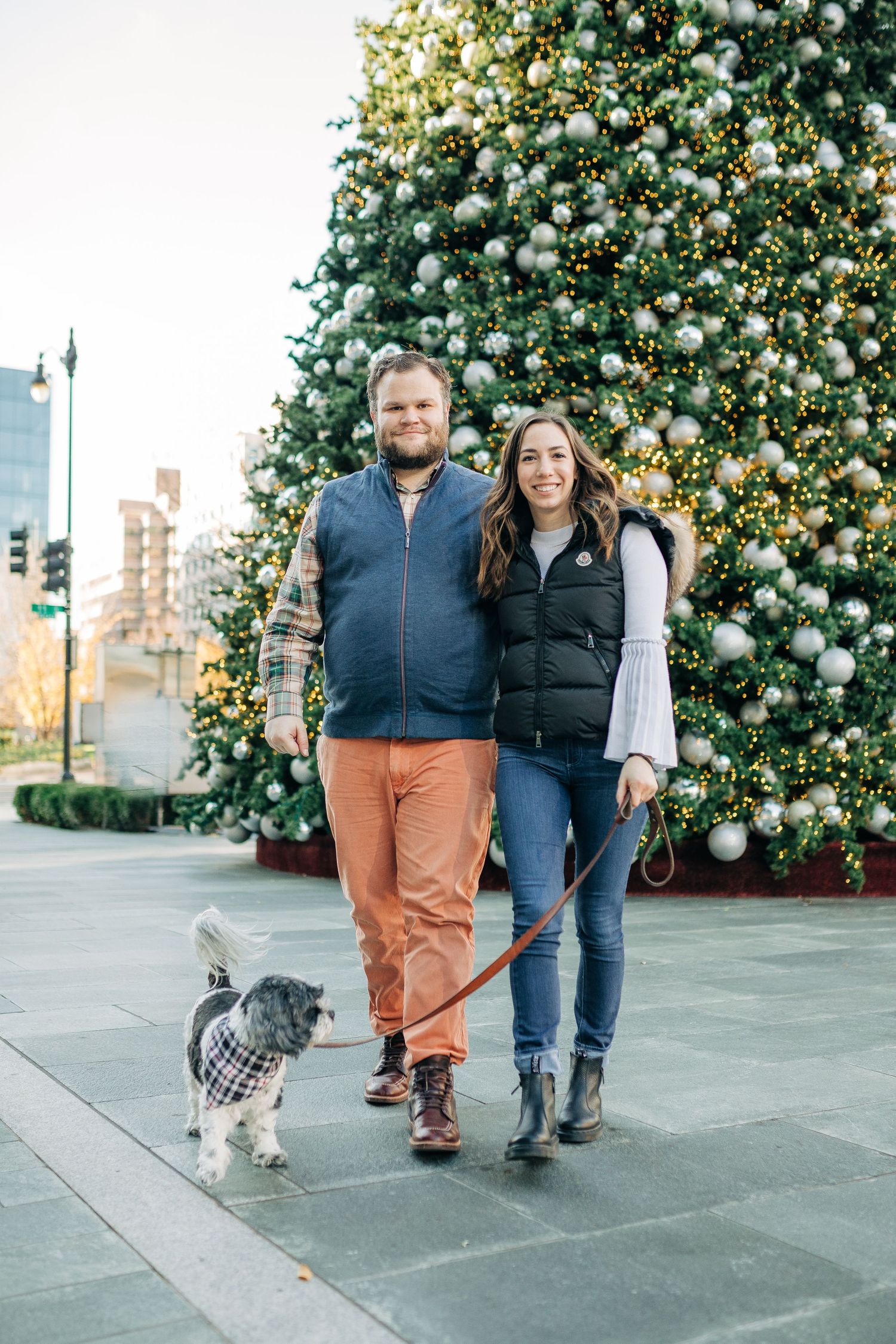 A couple walks their dog in front of a large decorated Christmas tree in an urban setting.