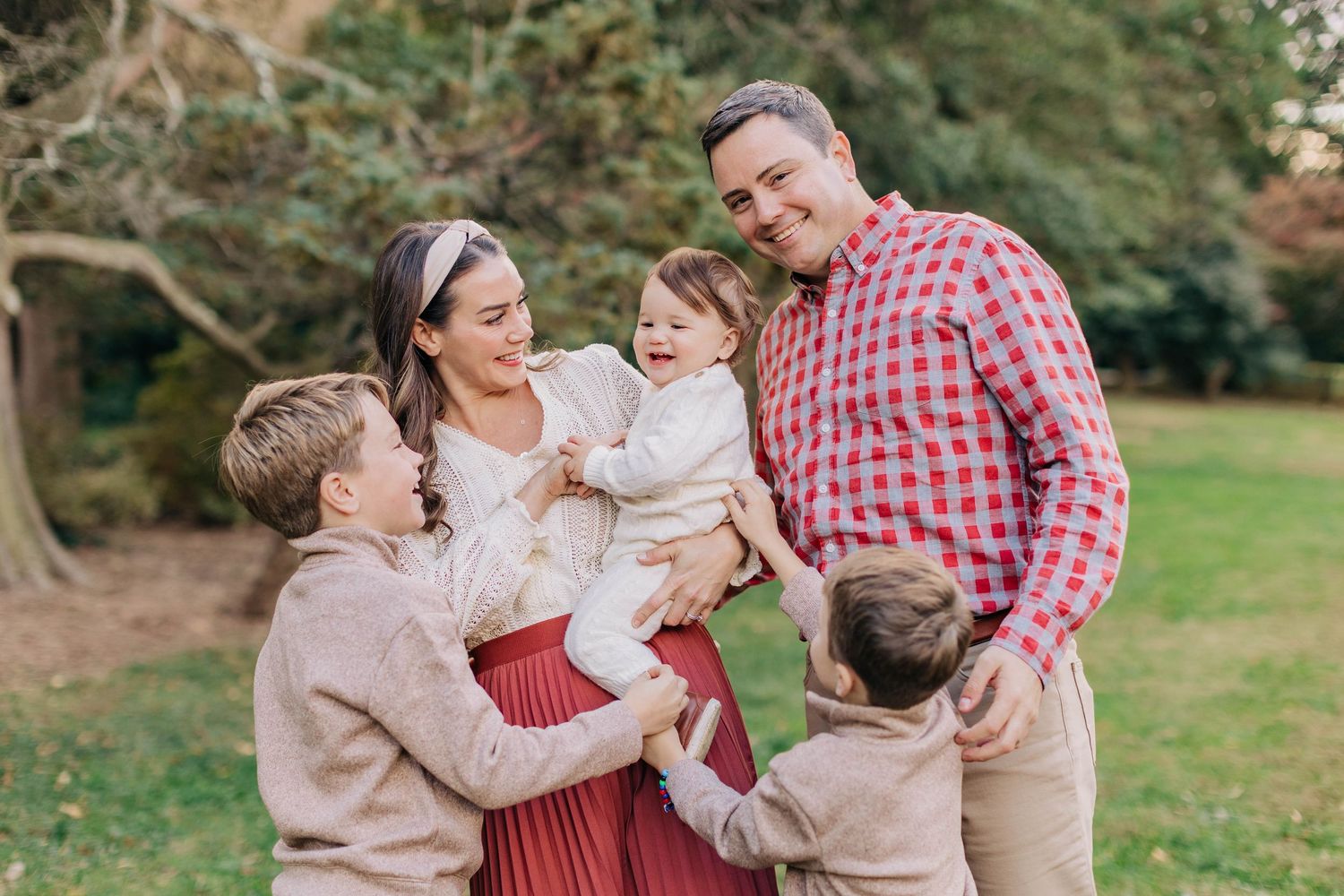 Happy family of five smiling outdoors in a lush green park, dressed in festive red and cream holiday attire.