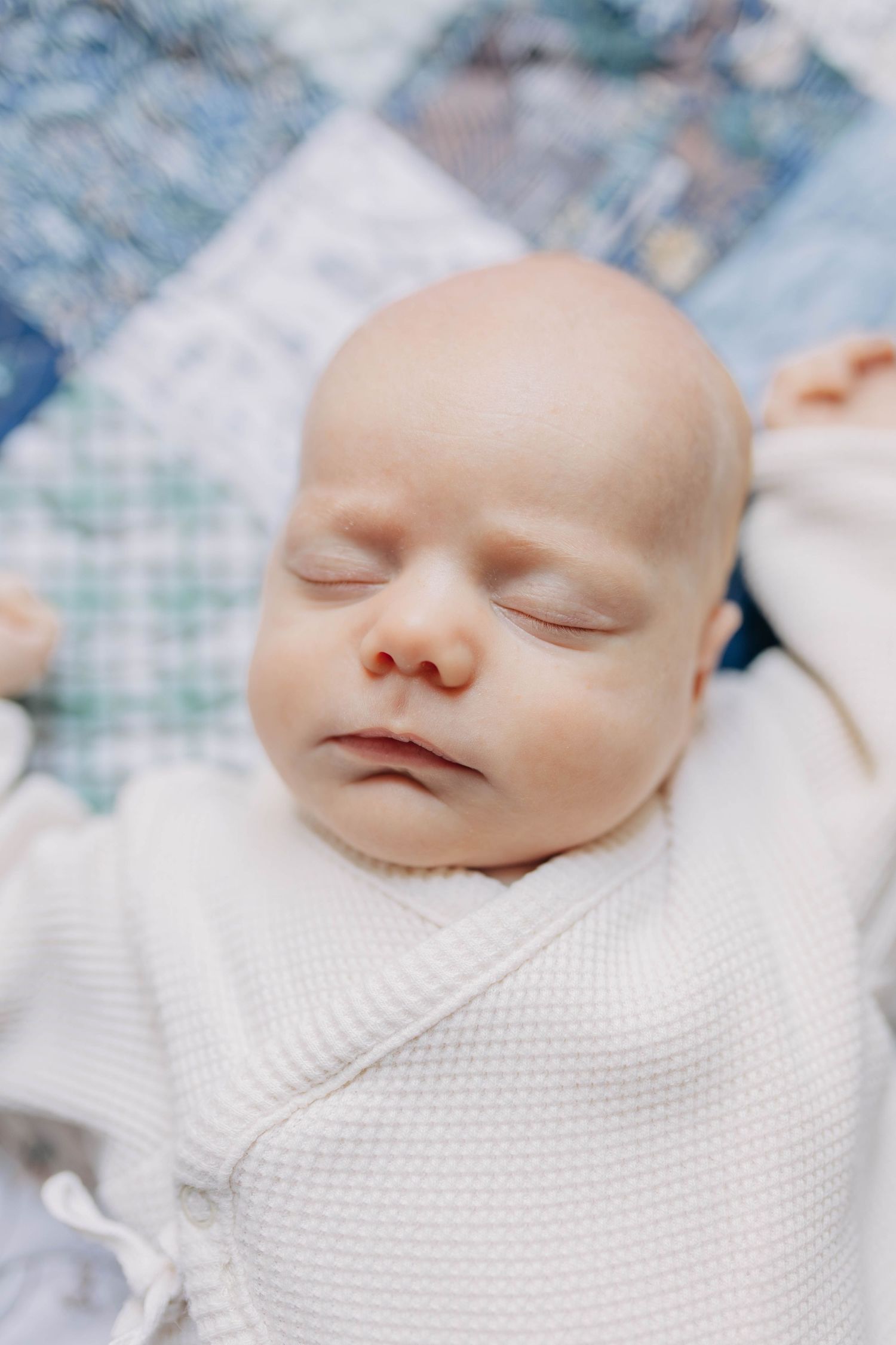 Peaceful sleeping newborn baby wrapped in white knit outfit lying on colorful patchwork quilt.