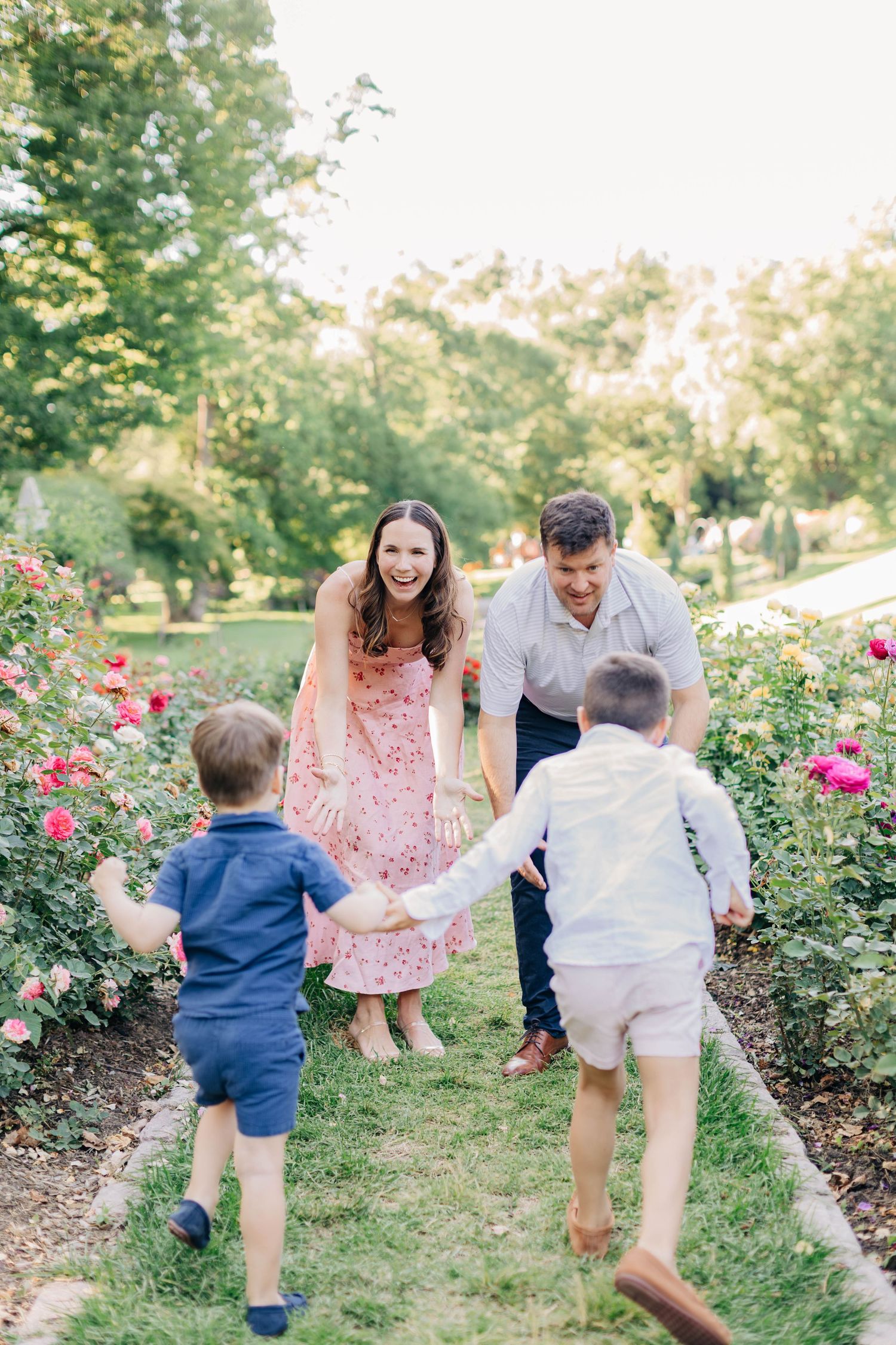 Family of four joyfully reuniting in a rose garden path, parents welcoming two young sons running toward them.