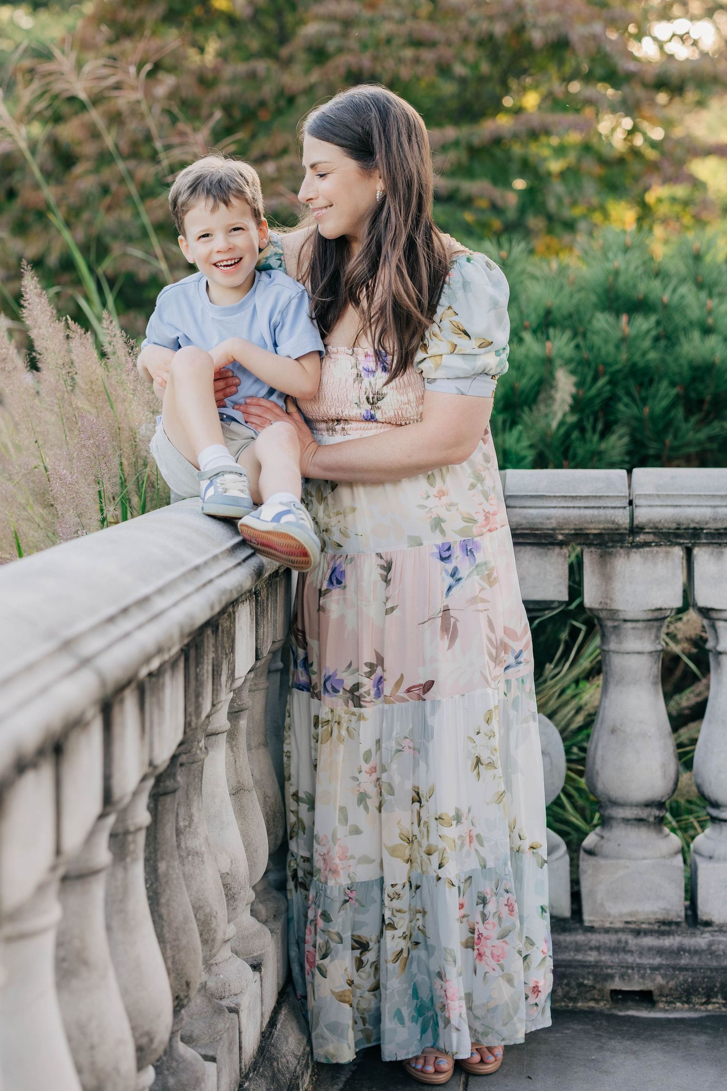 A smiling mother in a floral maxi dress holds her laughing son on a stone railing.