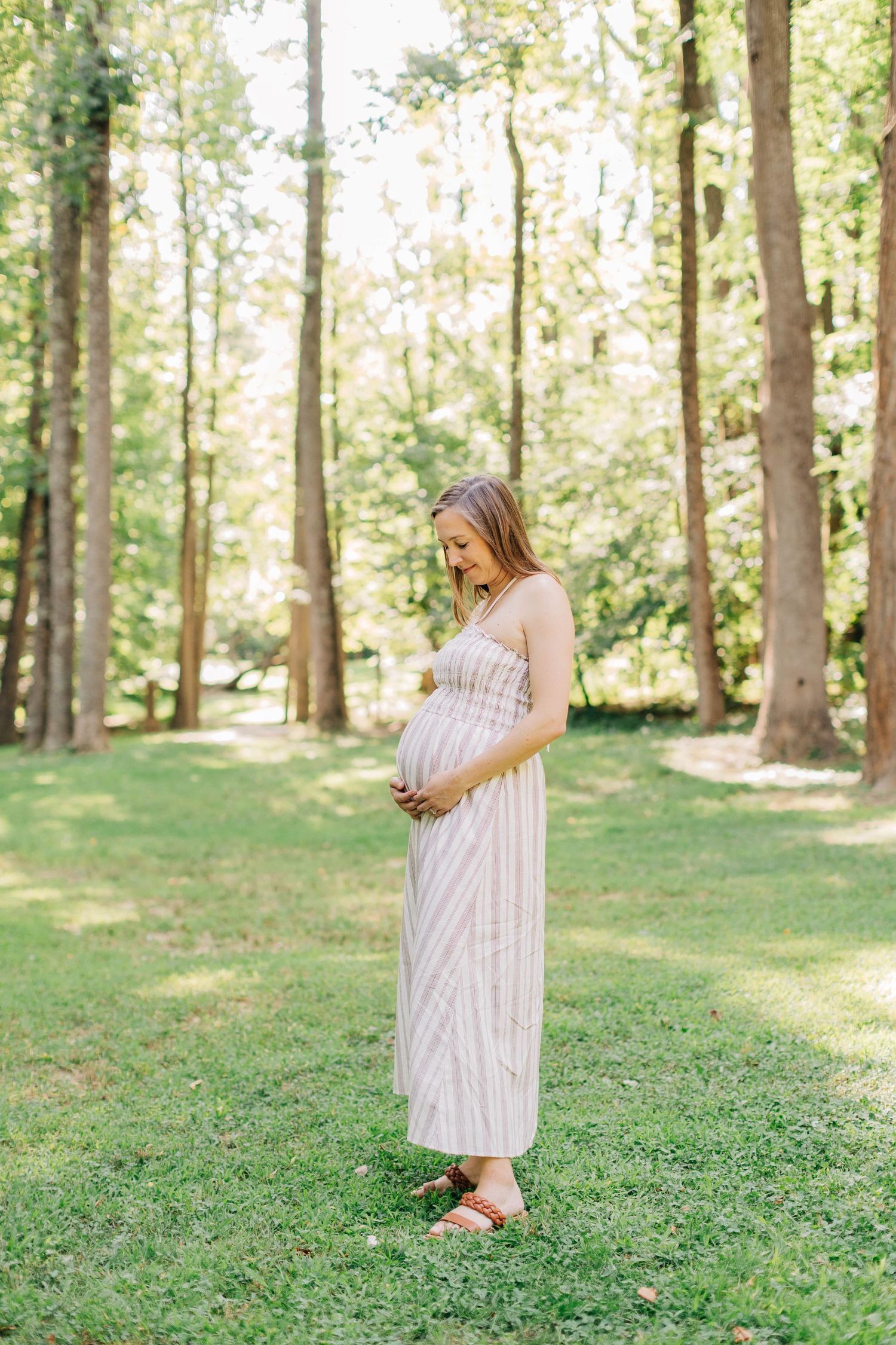 Pregnant woman in striped maxi dress standing on green grass in sunlit forest, cradling baby bump during maternity photoshoot.