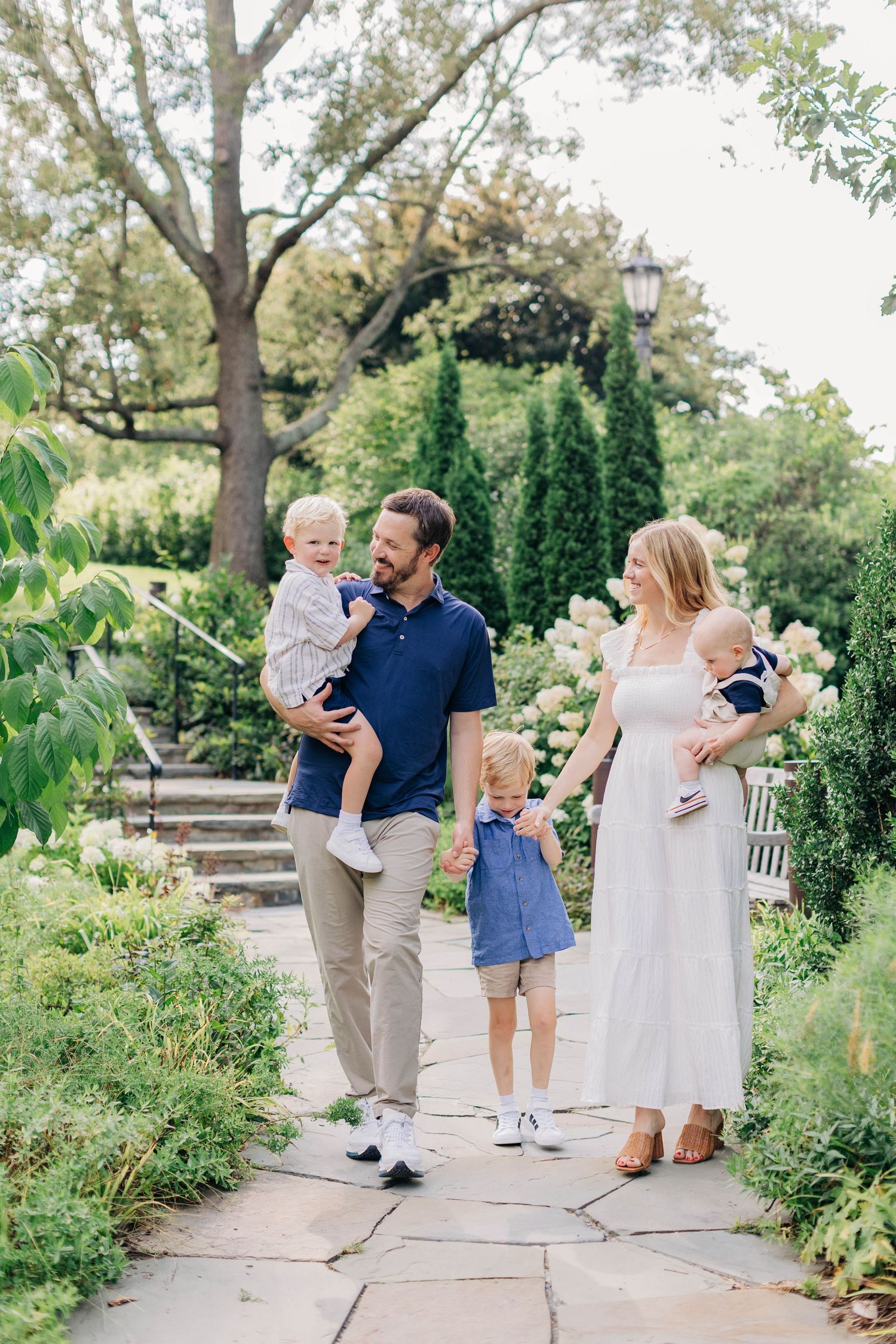 A happy family of five walks through a lush garden path, with parents holding toddlers and a young child between them.