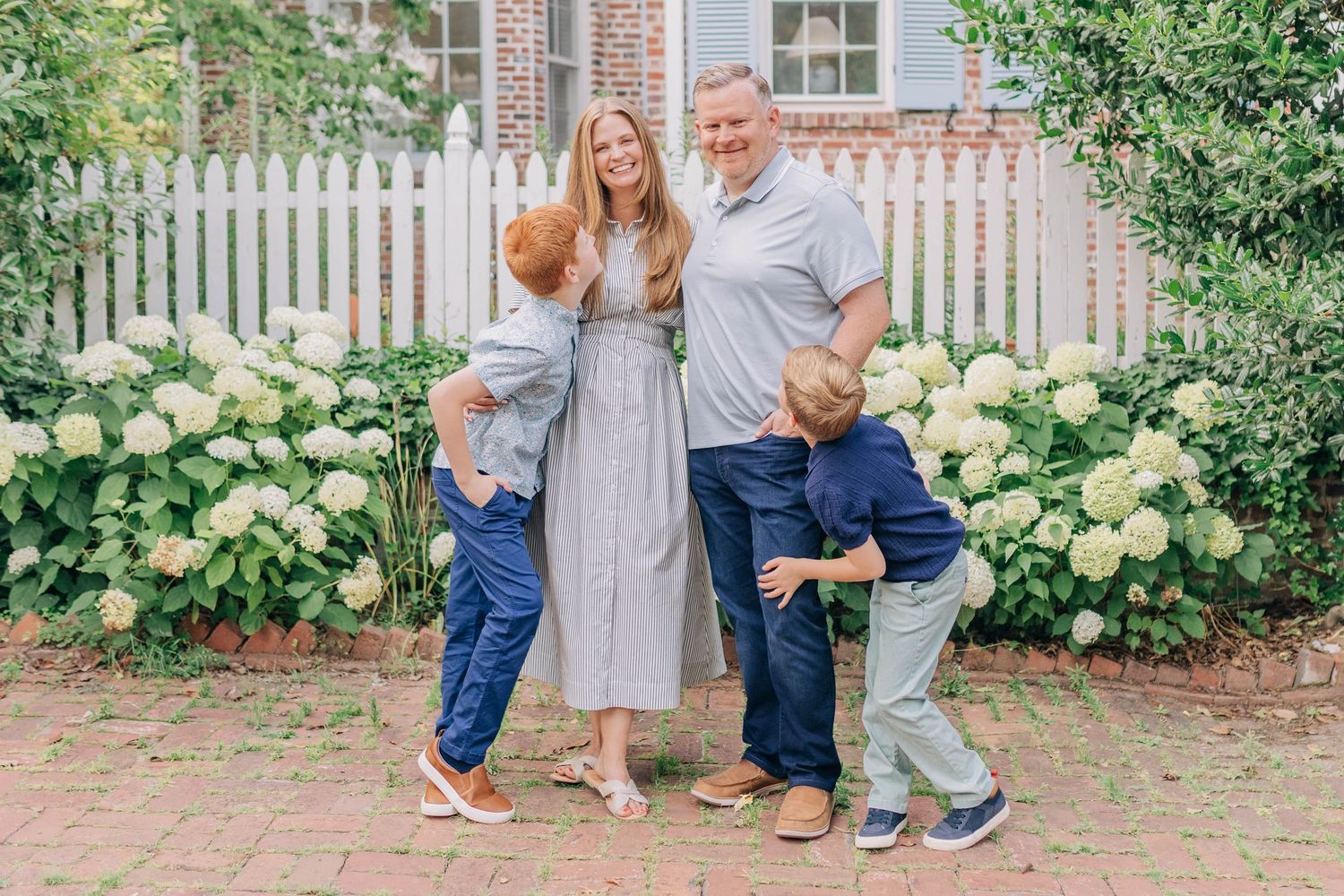 Family of four posing outdoors near white picket fence with blooming hydrangeas on brick pathway.
