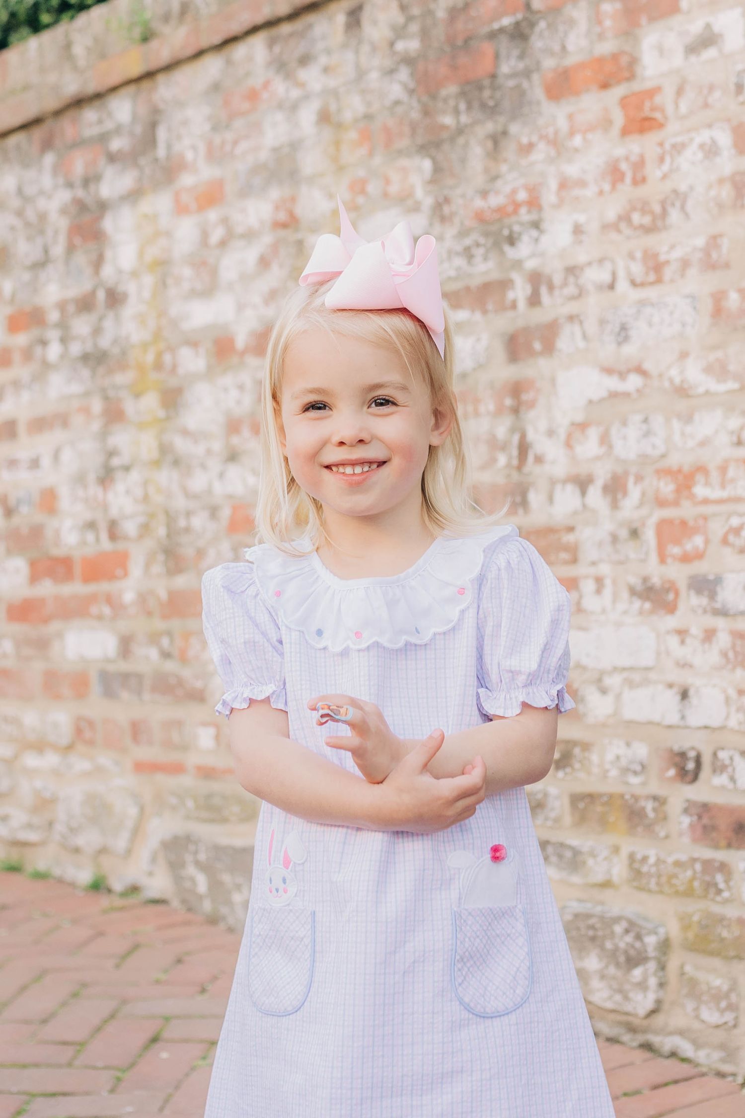 Young girl in lavender seersucker dress with pink bow, smiling in front of brick wall.