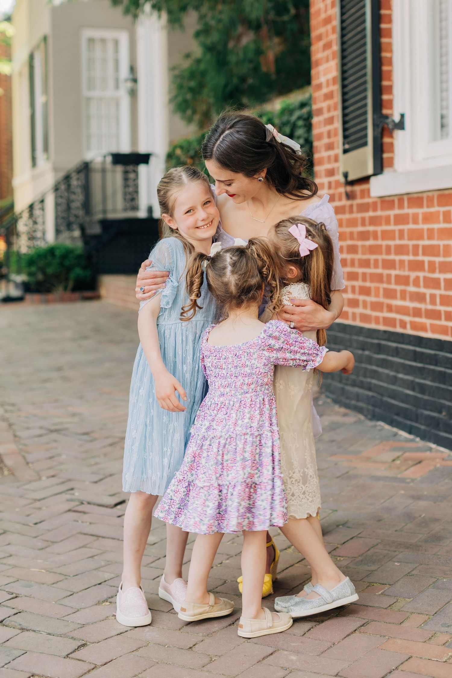 A mother hugging three young daughters in colorful dresses on a brick sidewalk outside a red brick building.