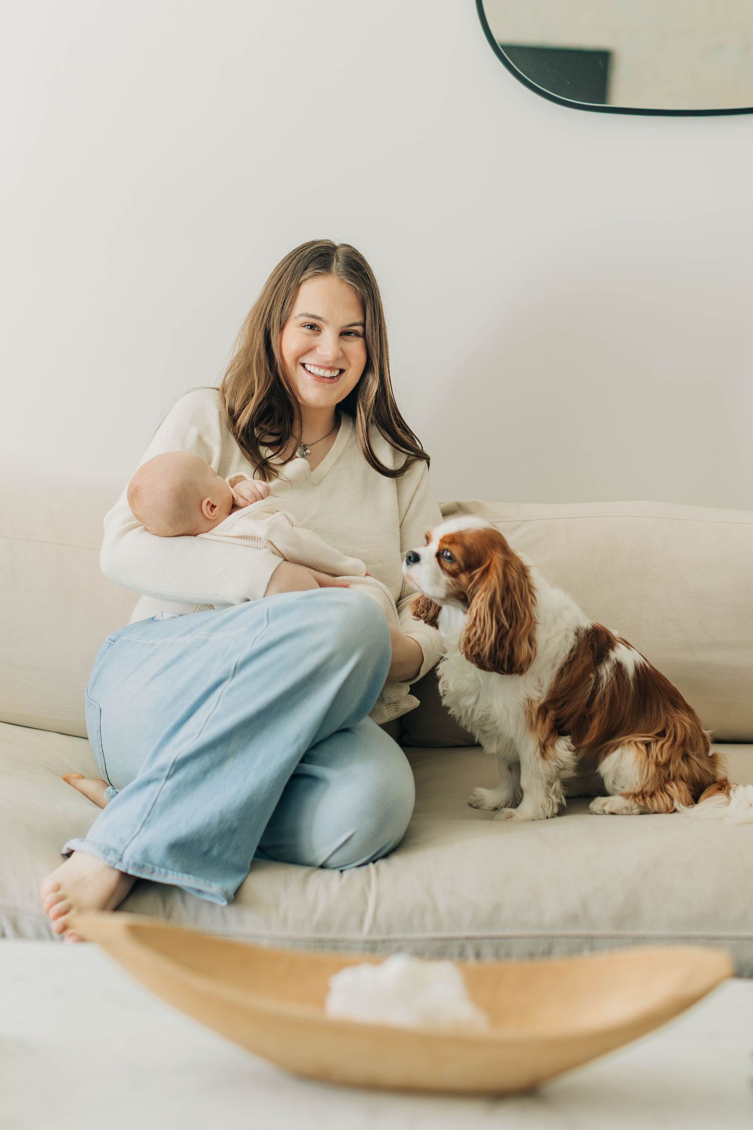 Smiling mother holding newborn baby on beige sofa with Cavalier King Charles Spaniel dog in bright living room.
