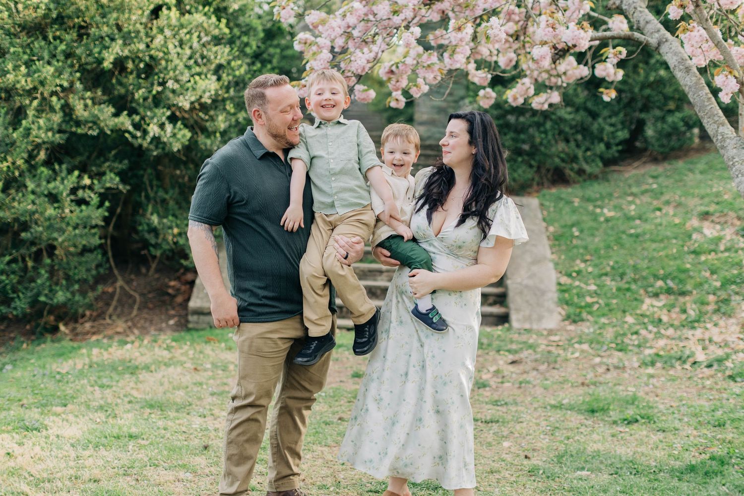 Happy family with two young boys posing under a blooming cherry blossom tree in a lush green garden.