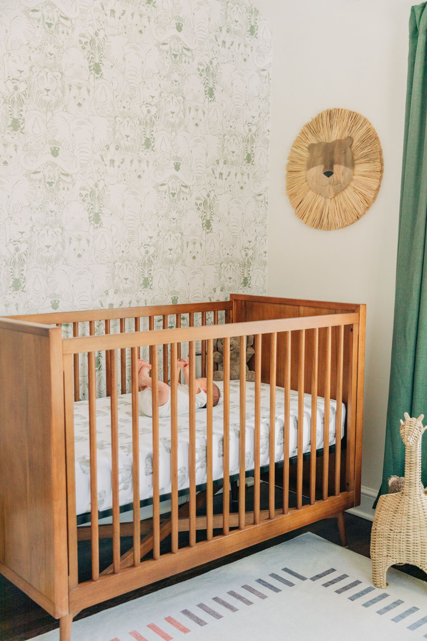 Wooden baby crib with striped rug in nursery room decorated with green curtains and wall decor.