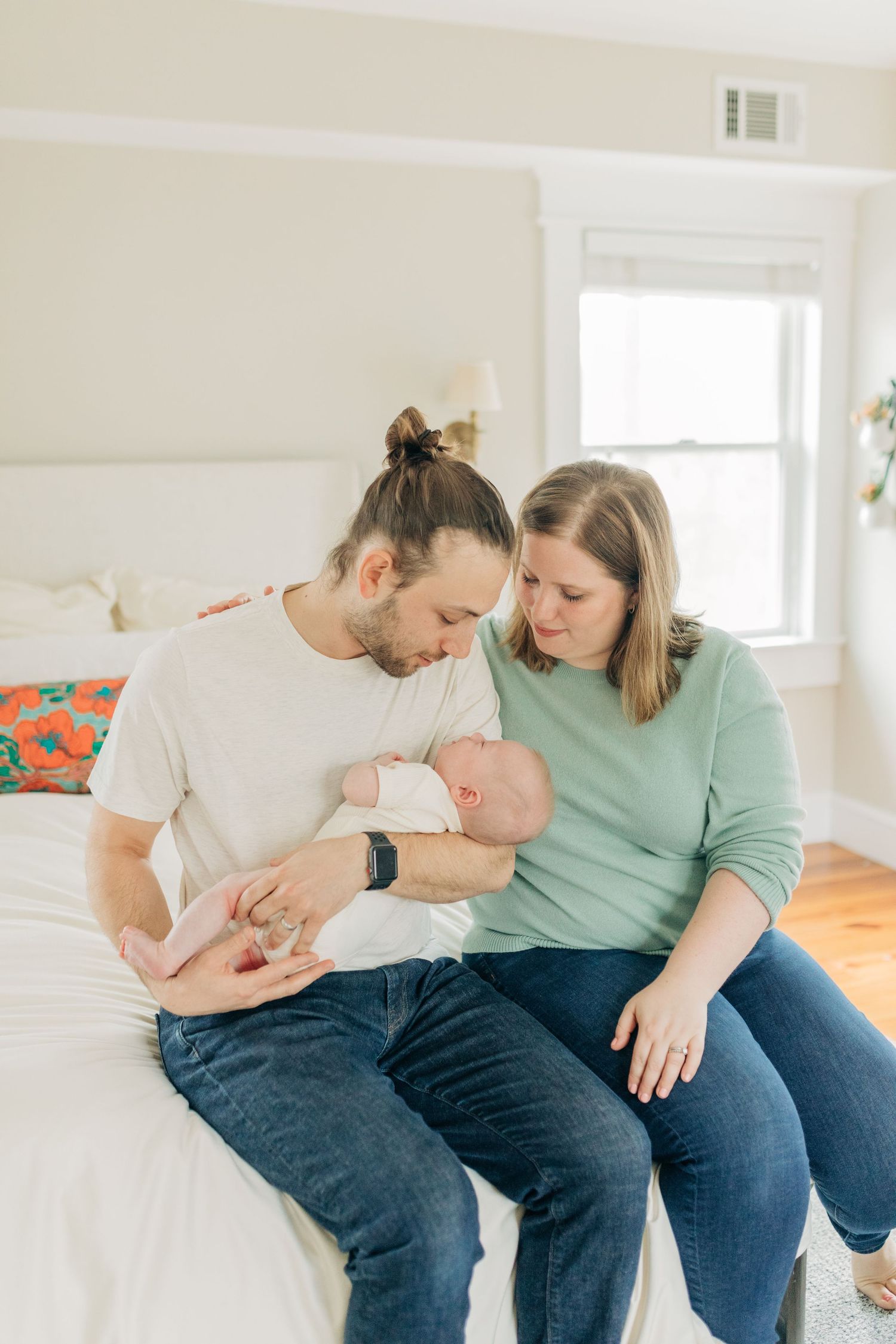Couple in jeans and casual wear sharing tender moments with their baby on a white bed.