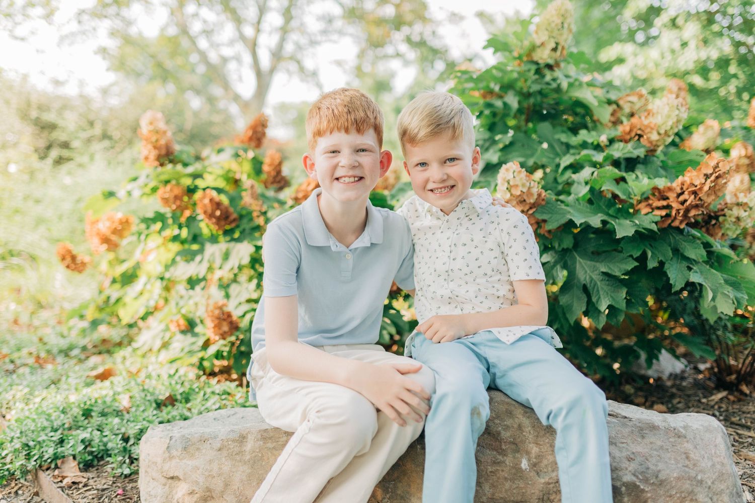 Two children sit together on a large rock in a garden setting with lush greenery in the background.