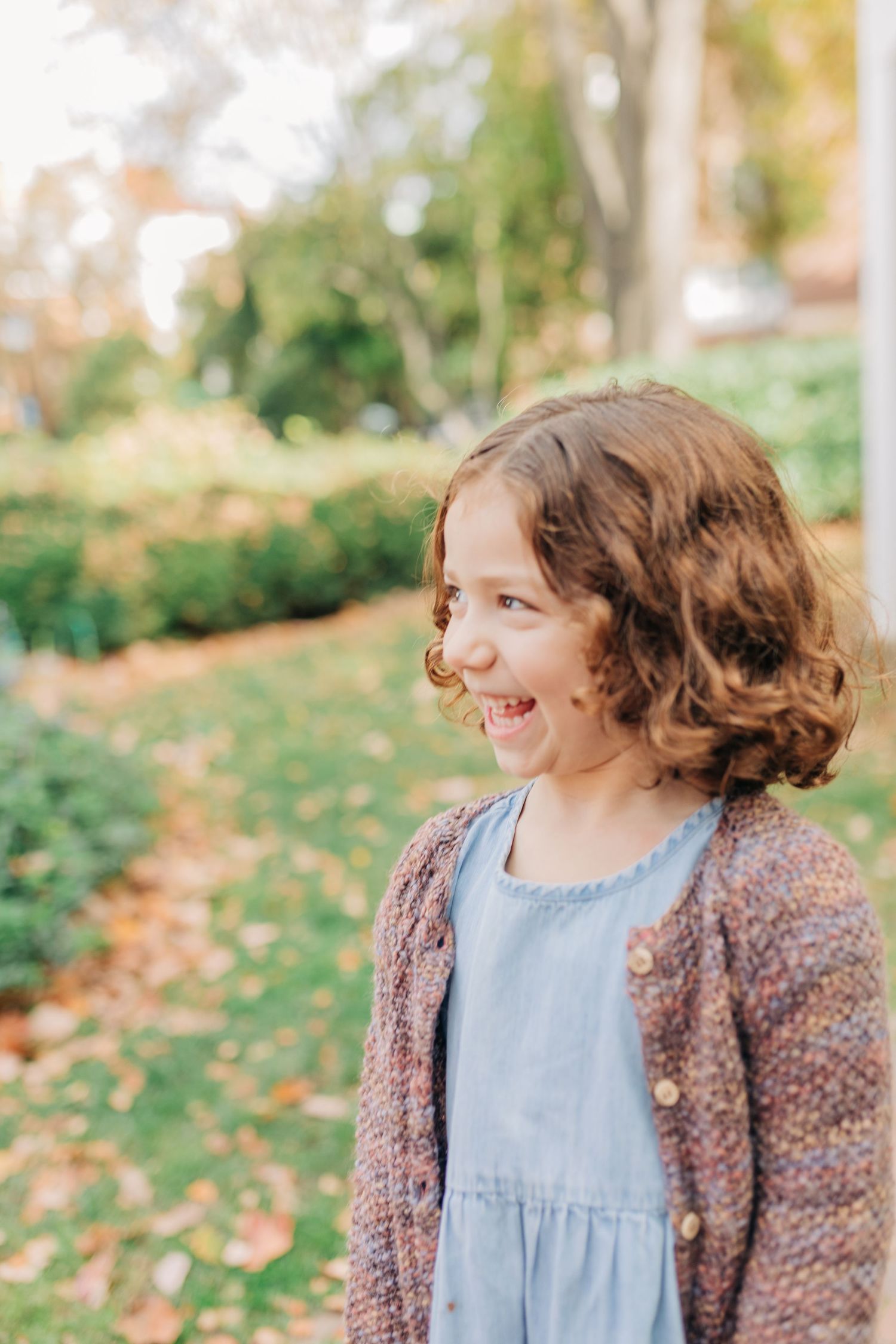 A cheerful young girl with curly hair smiles outdoors in a blue dress and knit cardigan among autumn leaves.