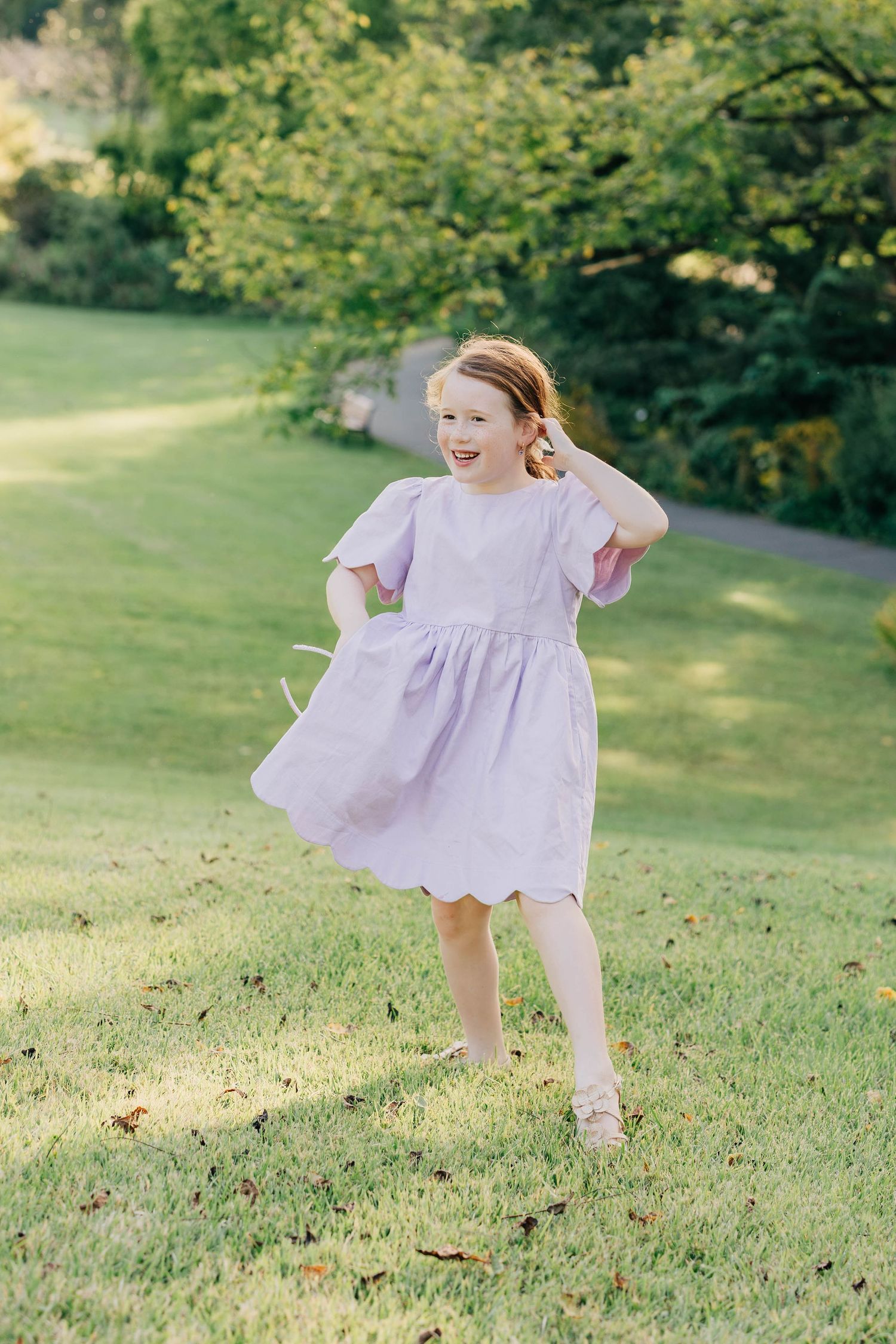 A joyful young girl twirls in a lavender scallop-hem dress on a sunny green lawn surrounded by lush trees.