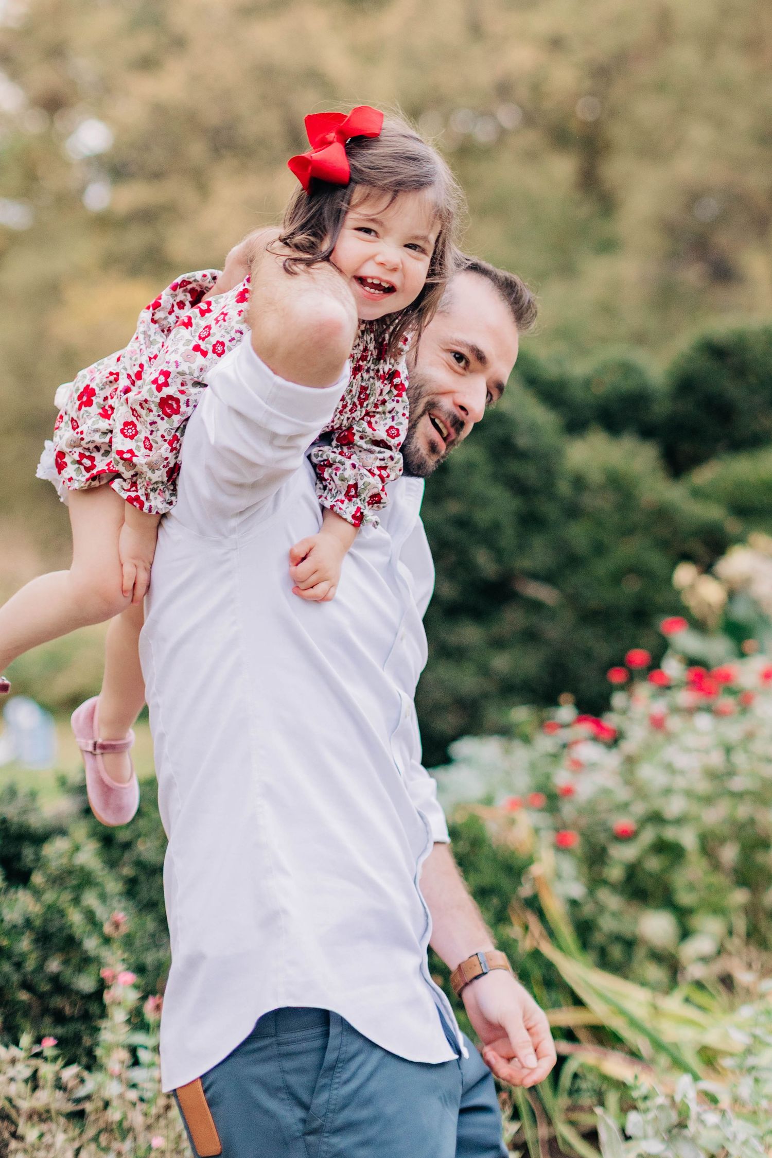Father joyfully holds his laughing daughter on his shoulder in a lush garden, girl wearing red bow and floral dress.