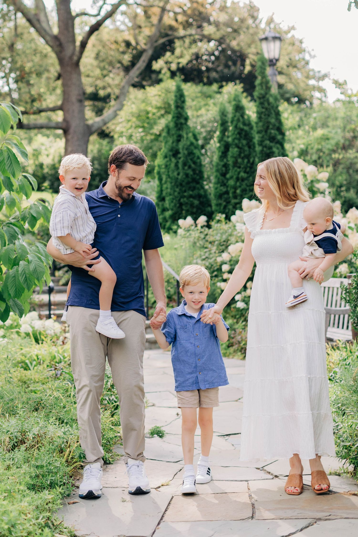 A happy family of five walks through a lush garden, parents holding children in coordinating blue and white outfits.