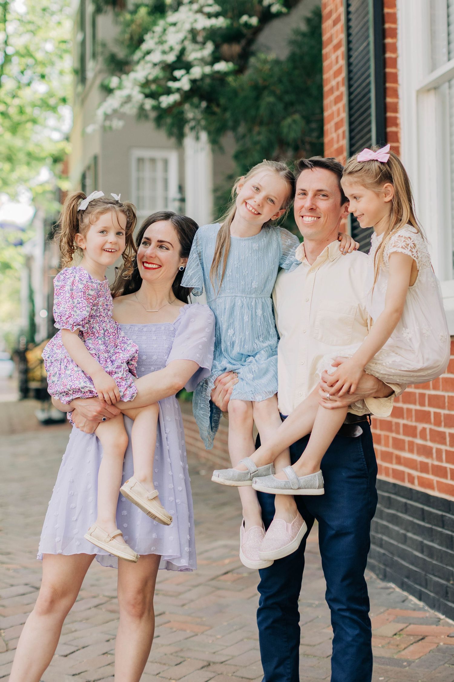 Happy family of five posing on a charming brick sidewalk, parents holding three young daughters in pastel outfits.