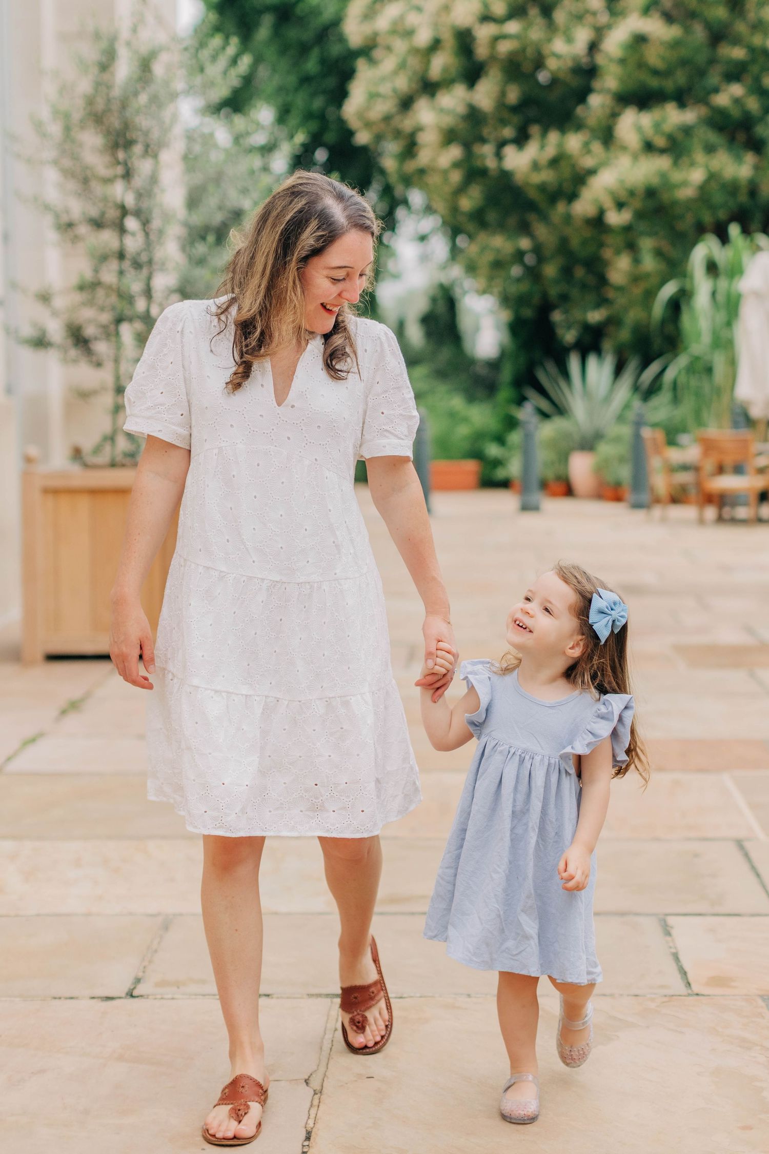 A smiling mother in a white dress holds hands with her young daughter in a blue dress while walking through a garden.